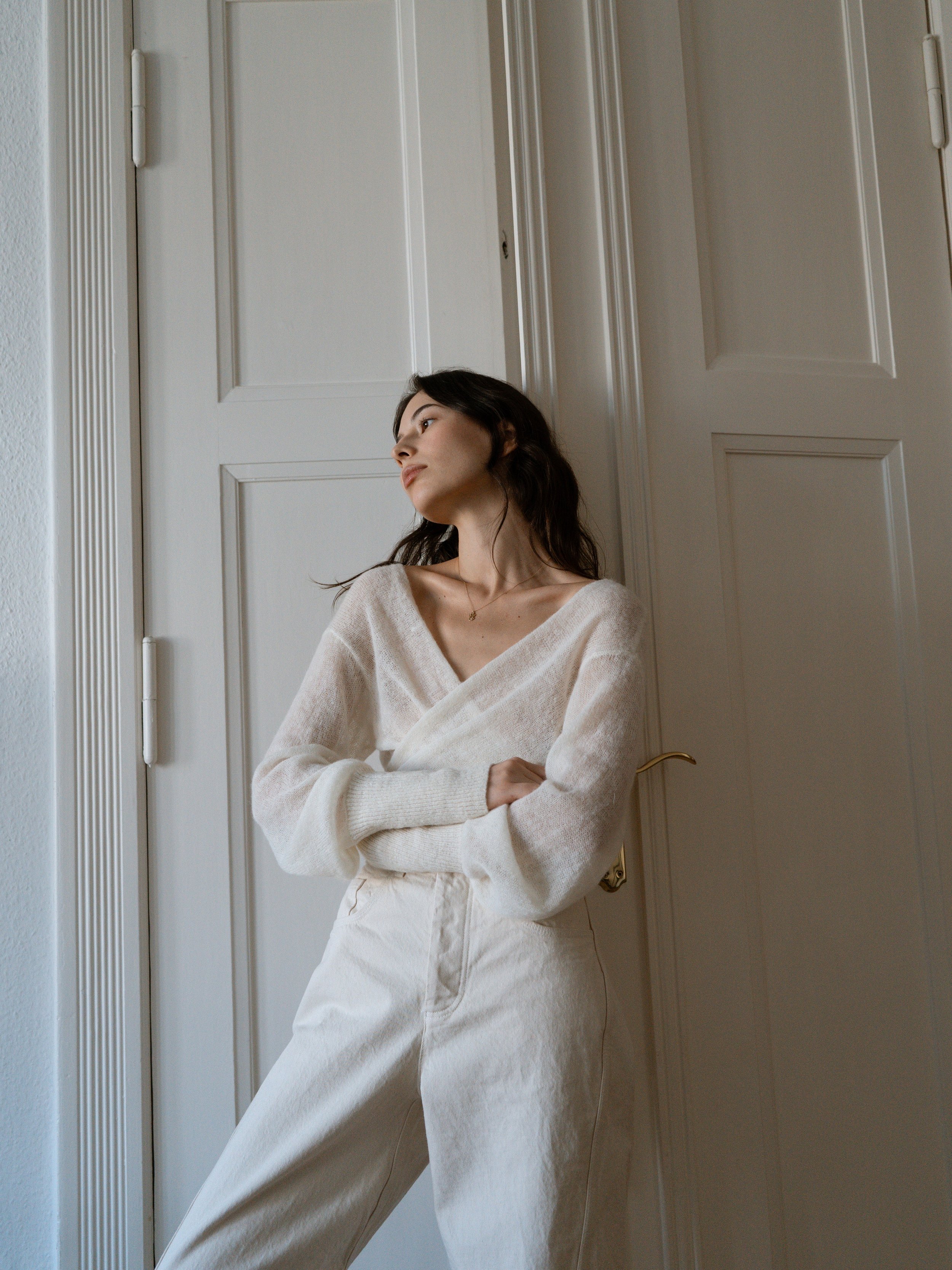 Woman in a white outfit standing against a wooden door, looking to the left with arms crossed.