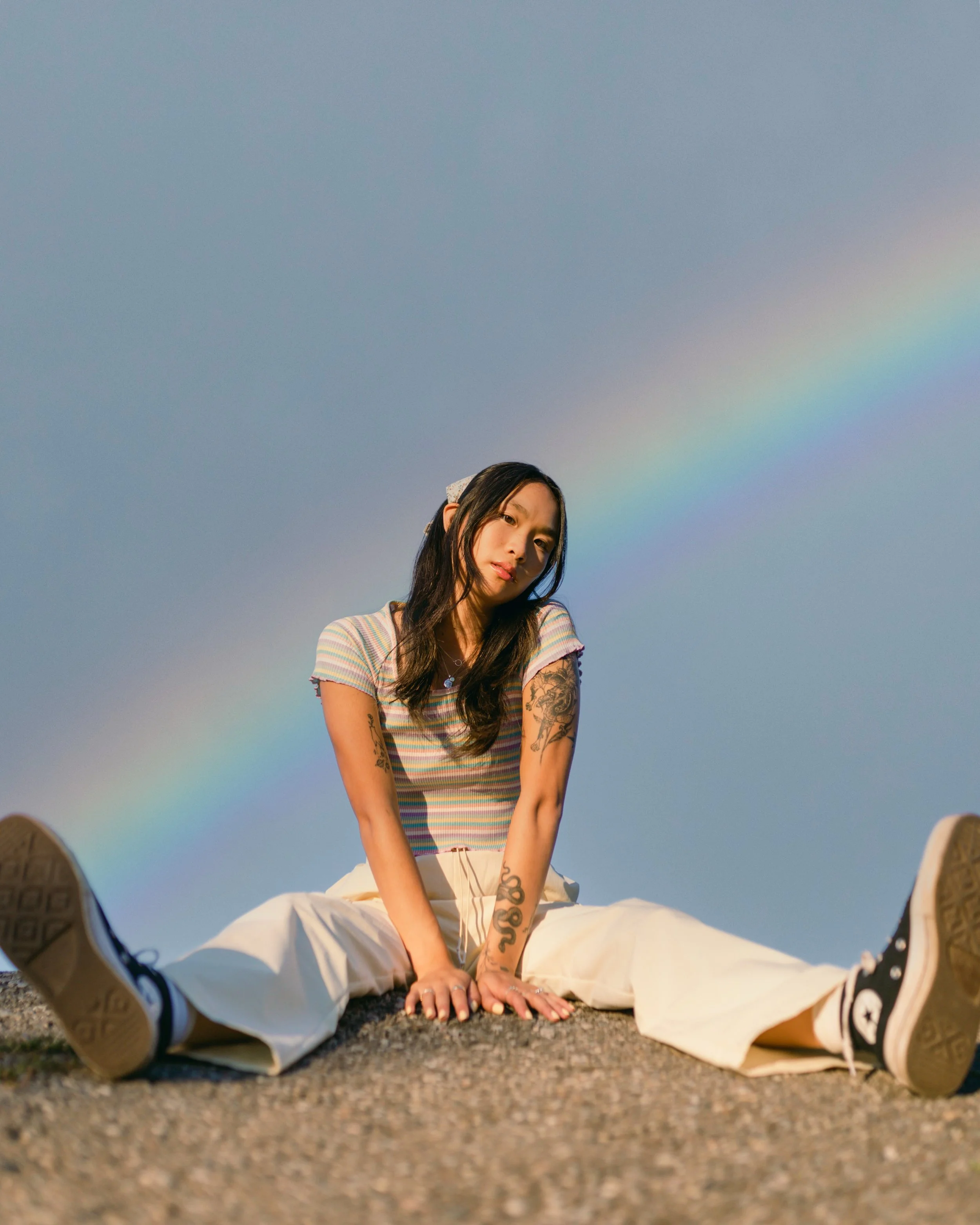 Person sitting on a surface with legs apart, wearing a striped shirt and white pants, with a rainbow in the background and tattoos visible on arms.