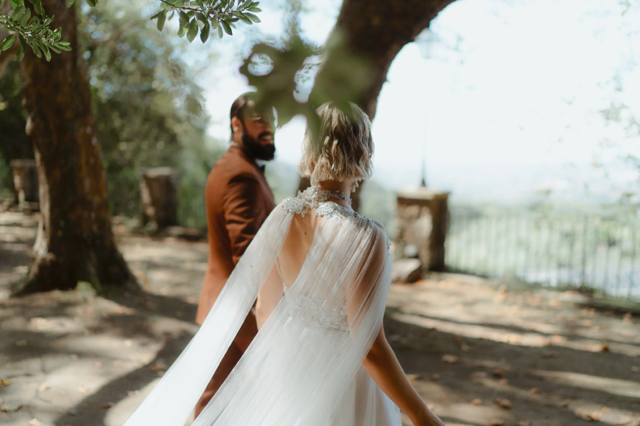 Bride and groom walking in a wooded outdoor setting, woman in wedding dress with flowing cape, tree branches overhead.