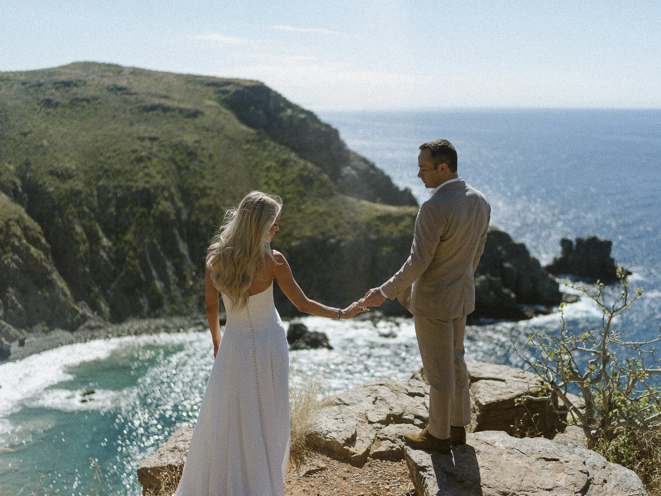 A couple holding hands on a rocky ledge overlooking the ocean, with cliffs and hills in the background, during daytime.