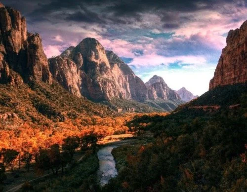 Scenic view of towering mountains with a river winding through a lush valley in autumn colors under a cloudy sky.