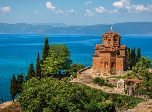 Church of St. John at Kaneo overlooking Lake Ohrid, surrounded by trees and greenery, with mountains in the background.
