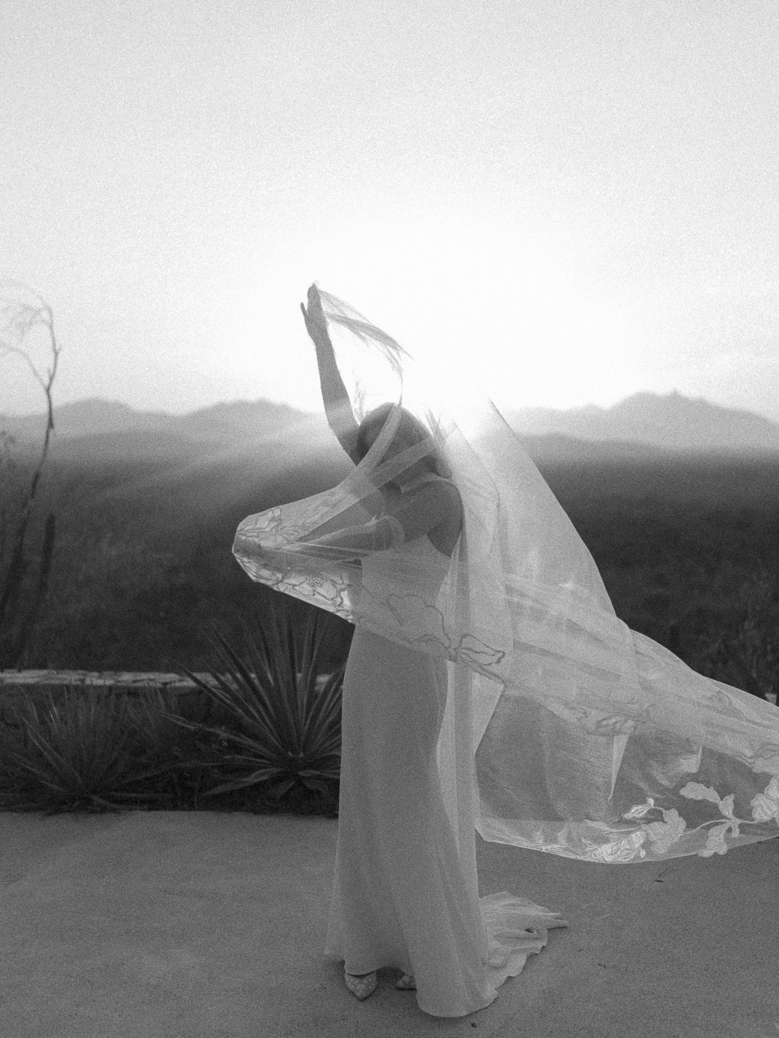 A woman dressed in a flowing, sheer dress holding a large piece of fabric or veil in an outdoor desert landscape at sunset or sunrise.