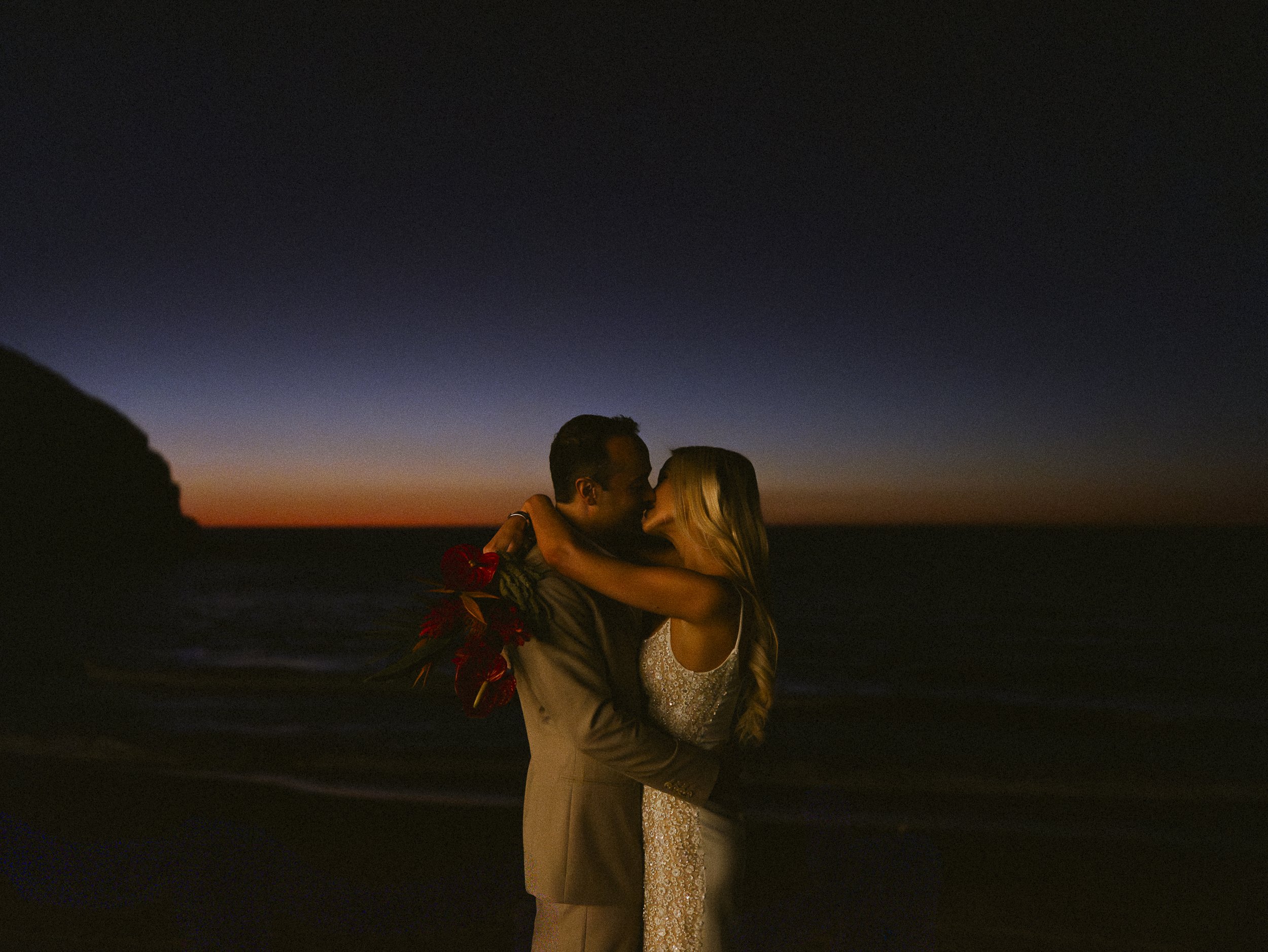 A couple embracing and kissing at the beach during sunset, with a colorful sky and ocean in the background.