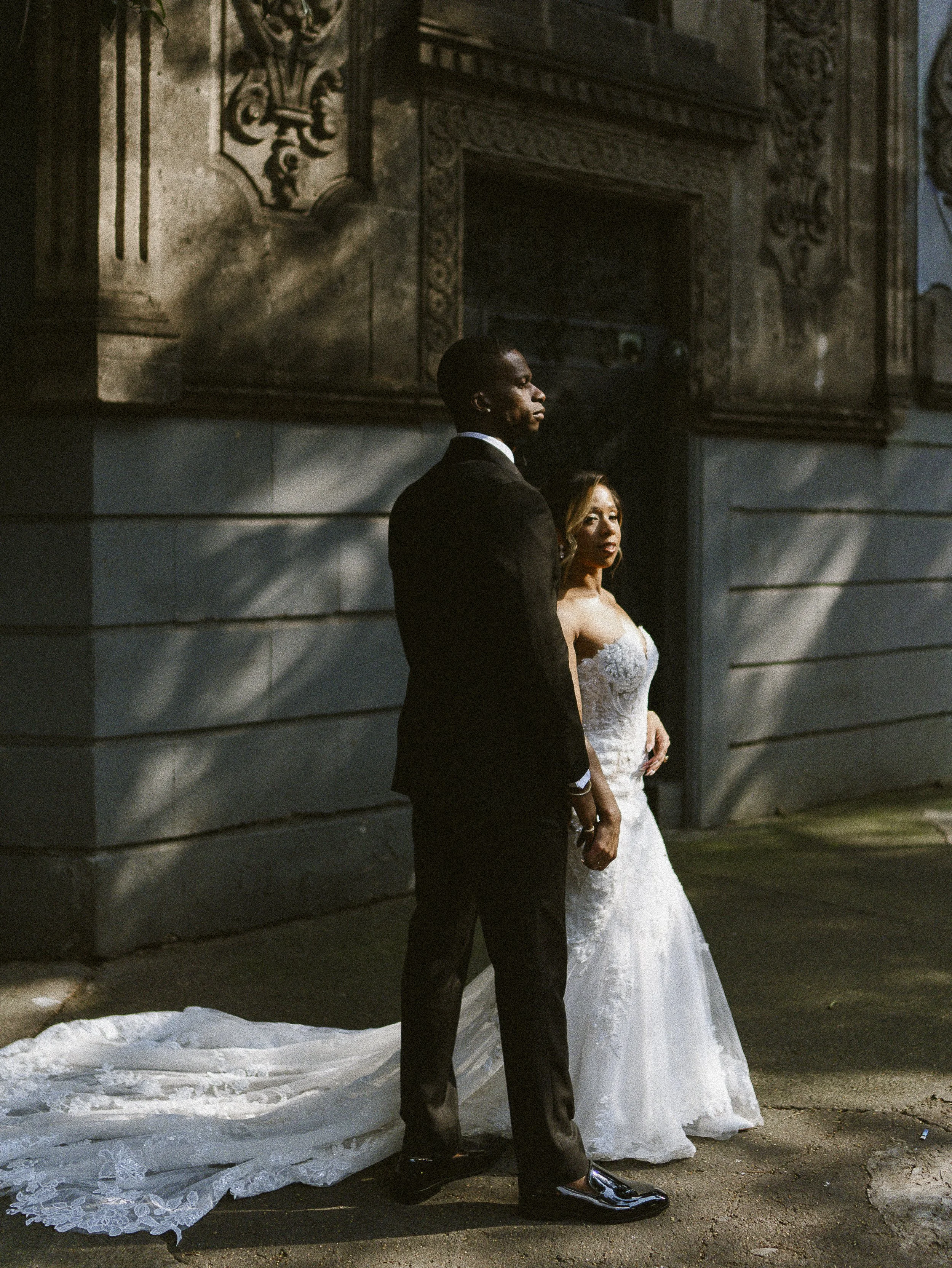 A bride in a white wedding gown and a groom in a black tuxedo stand close together outside in front of a stone and wood building, with the bride looking to her right.