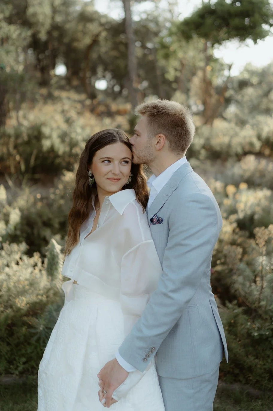 A couple embracing outdoors, with the man kissing the woman's forehead. The woman is wearing a white outfit and the man is in a light gray suit. They are surrounded by greenery.