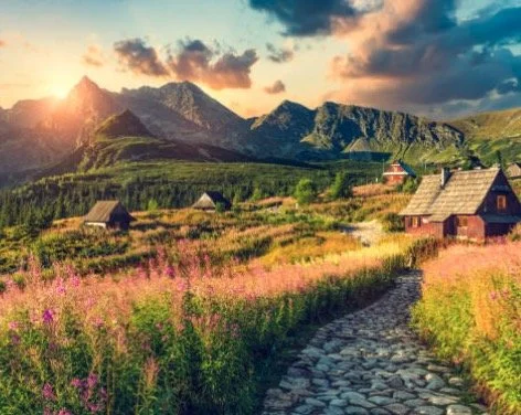 Scenic mountain landscape with a cobblestone path leading to rustic wooden cabins surrounded by fields of wildflowers, under a colorful sunset sky.