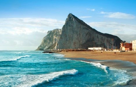 Rocky coastline with a prominent cliff, sandy beach, and ocean waves under a blue sky.