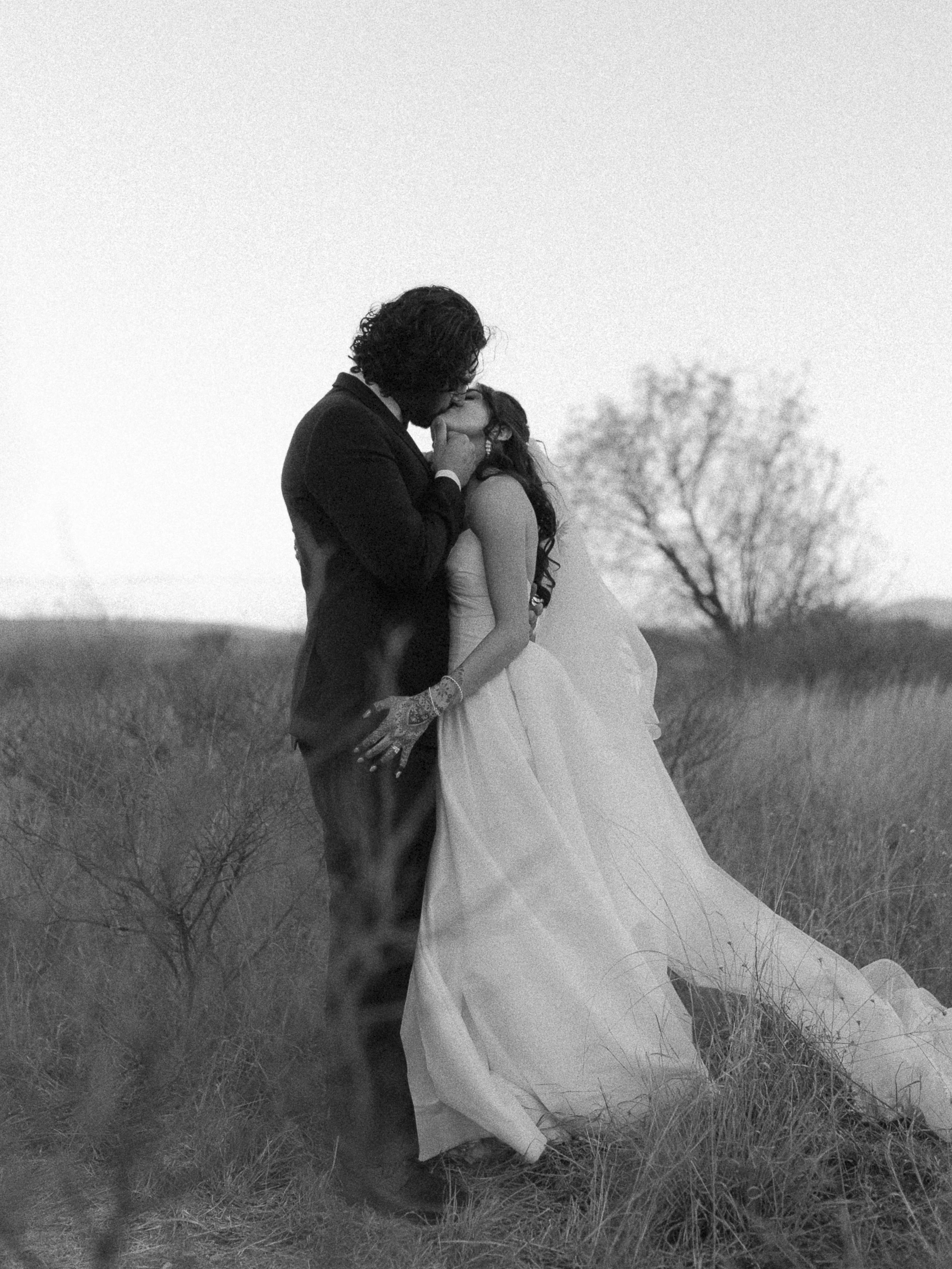 A black and white photo of a couple kissing in a field, with one wearing a wedding dress and the other in a suit.