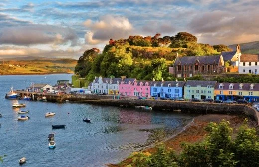 A scenic coastal village with colorful buildings lining the waterfront. Small boats float in the calm harbor. Lush greenery and trees surround the area, with hills in the background under a cloudy sky.