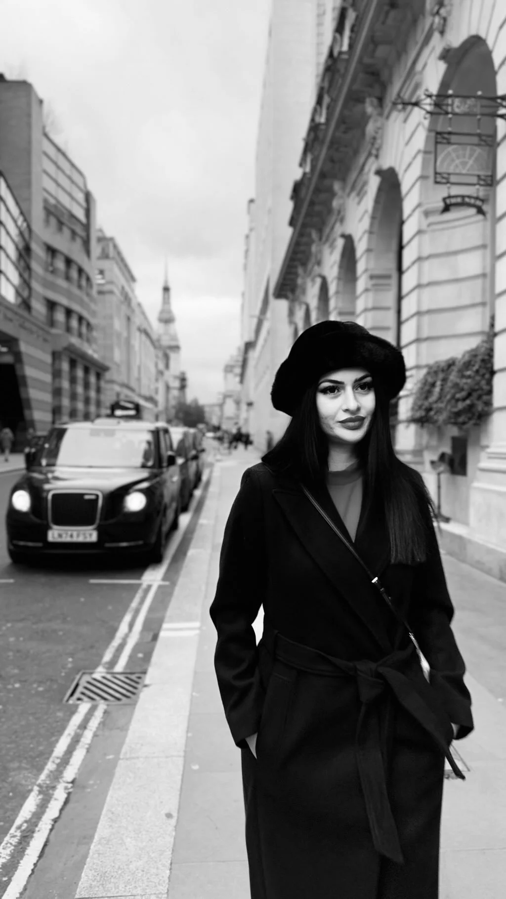 A woman dressed in black walking on a city street with European architecture, cars parked along the side, and a spire in the background.