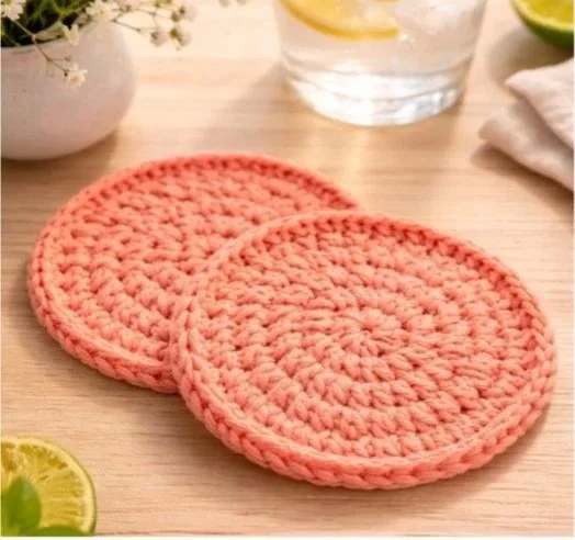 Two pink crocheted coasters on a light wooden table with a glass of water, a bowl of white flowers, and lime slices in the background.