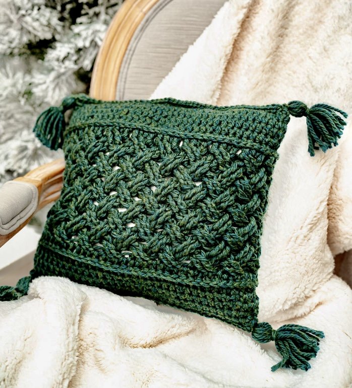 Green crocheted bag with tassels, resting on a beige fleece blanket, with a wooden chair arm and Christmas tree in the background.