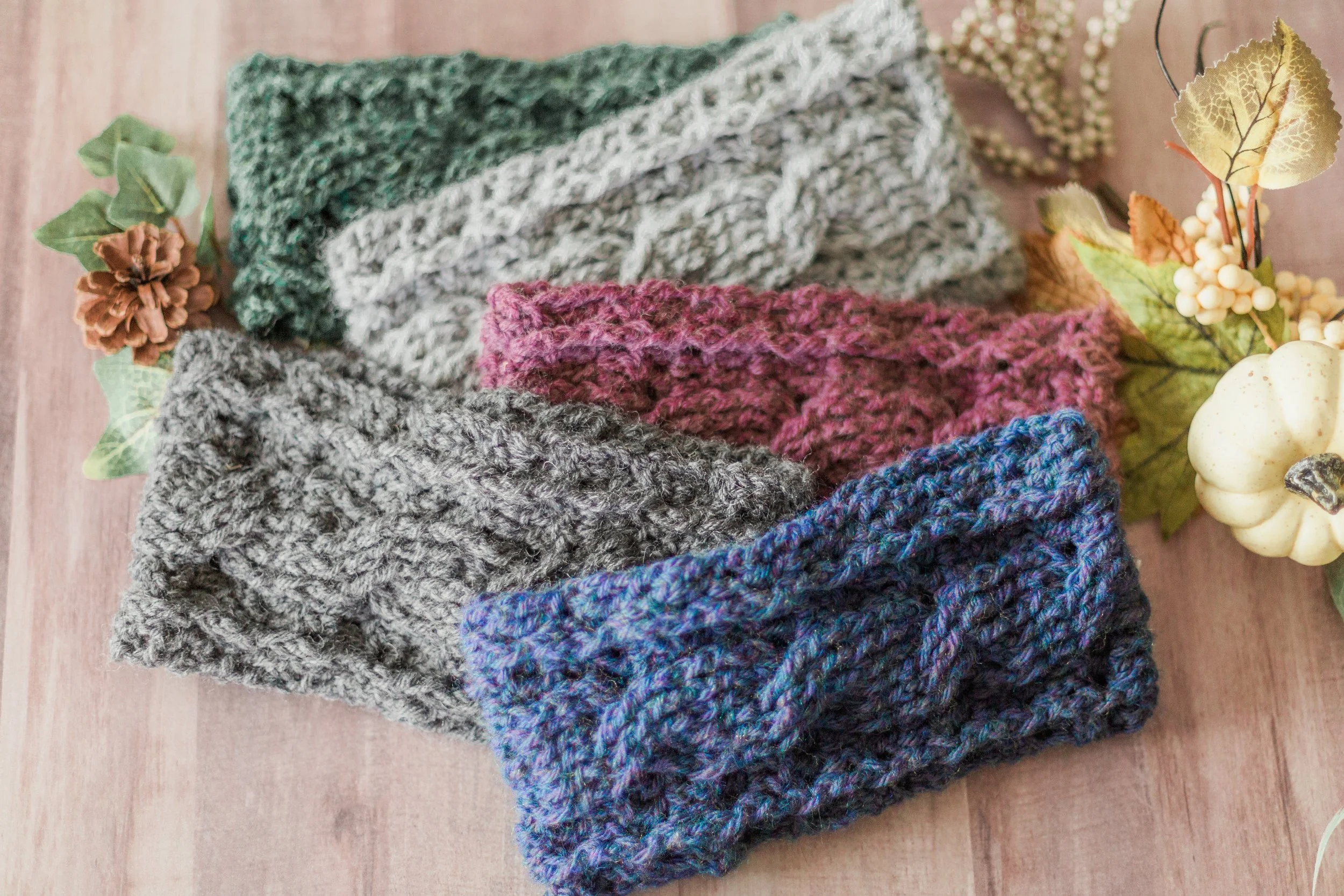 A collection of crochet ear warmersin various colors including gray, teal, pink, purple, and blue, arranged on a wooden surface, with decorative fall-themed items such as pinecones, faux pumpkins, and leaves around them.