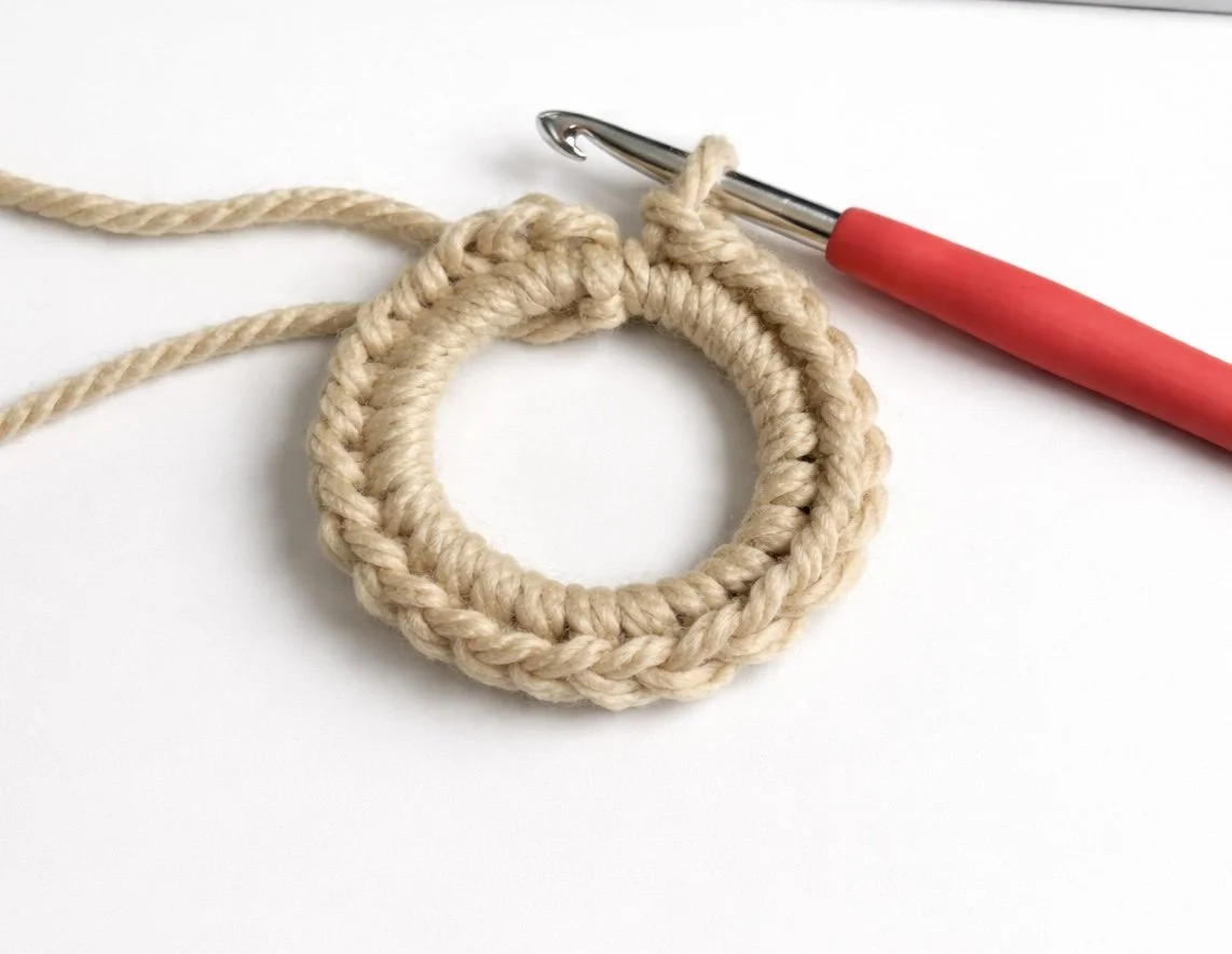A crochet project with beige yarn, a crochet hook with a red handle, and an unfinished circular yarn piece on a white surface.