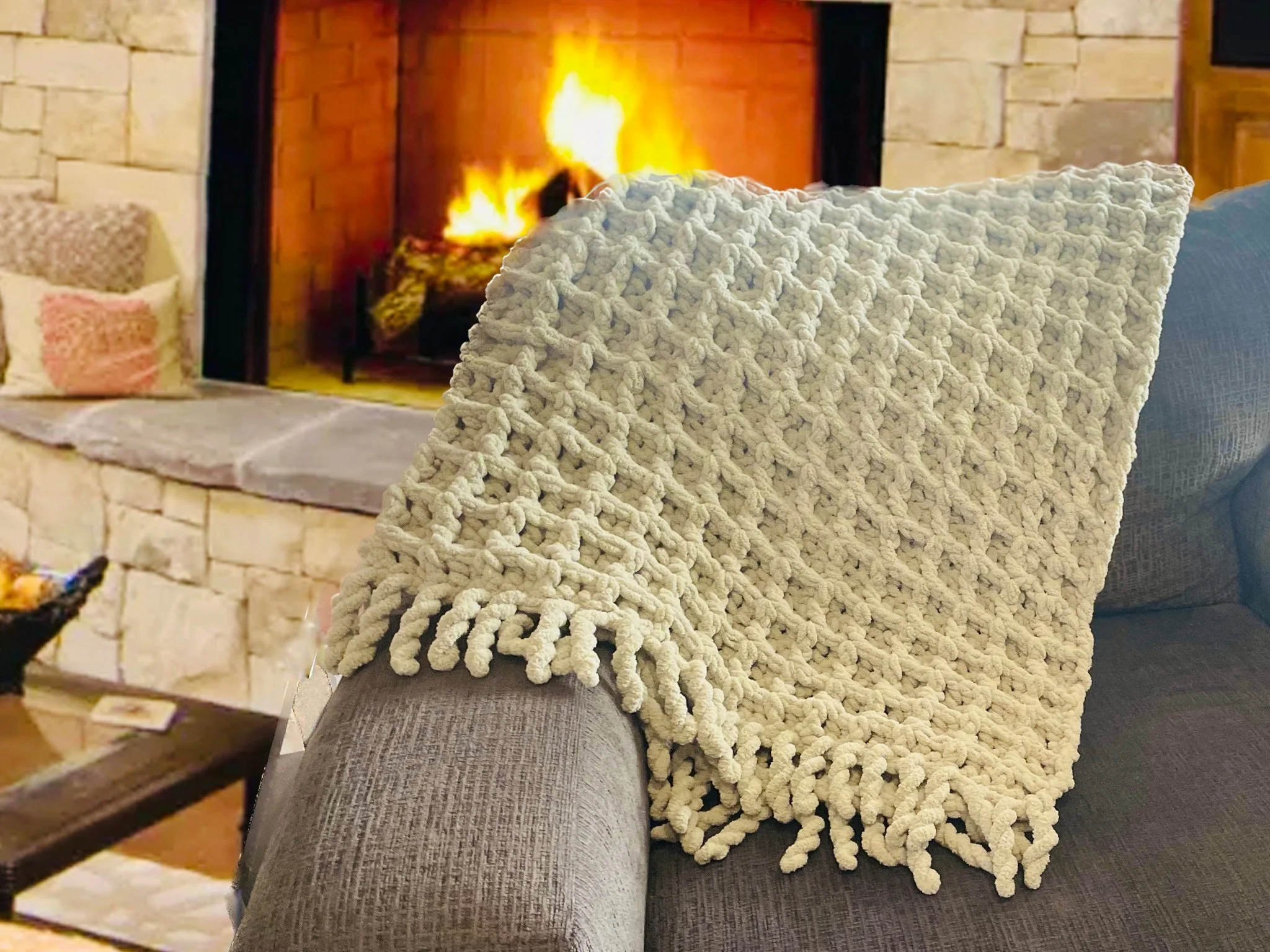 A cozy living room corner with a knitted cream-colored throw blanket draped over a gray armchair, a stone fireplace with a fire burning inside, and a side table with pillows nearby.