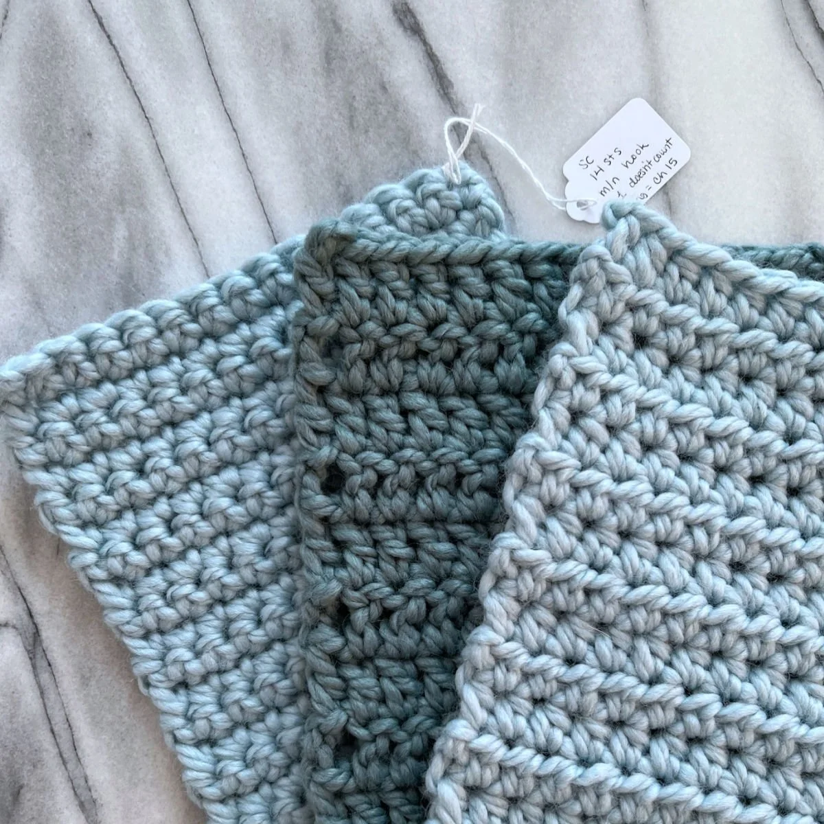 Close-up of knitted gray and white fabric swatches with a small handwritten tag attached, lying on a light-colored marble surface.
