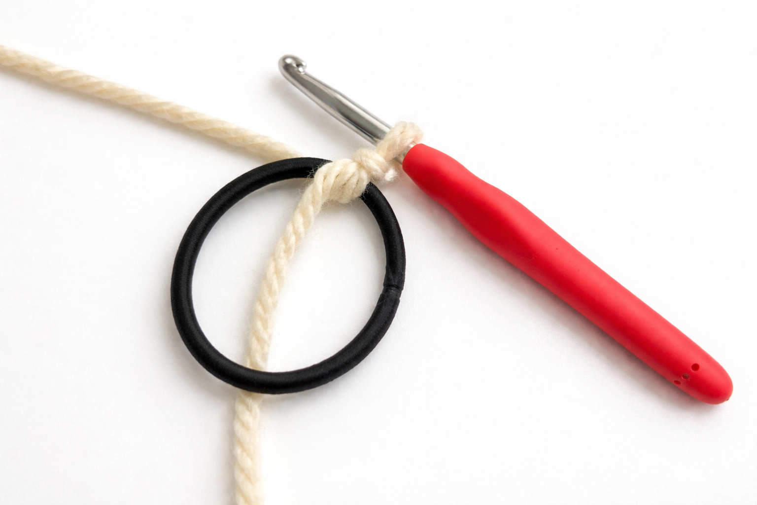 A crochet hook with a red handle, a black elastic hair tie, and cream colored yarn tied together, placed on a white background.