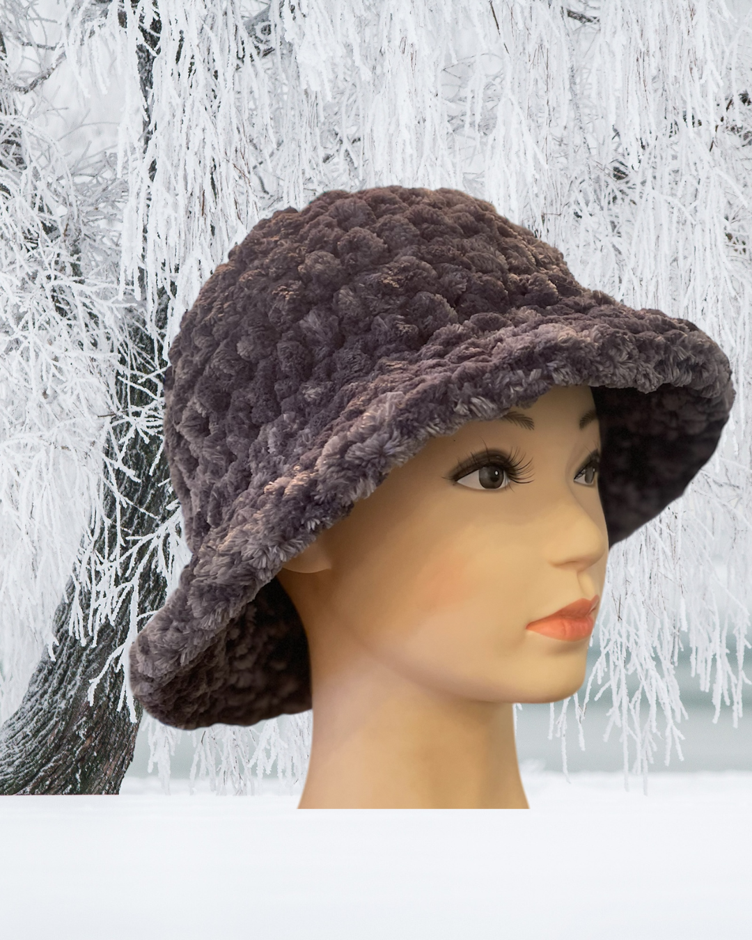 A mannequin head wearing a fuzzy, textured brown hat against a wintery background of snow-covered trees.