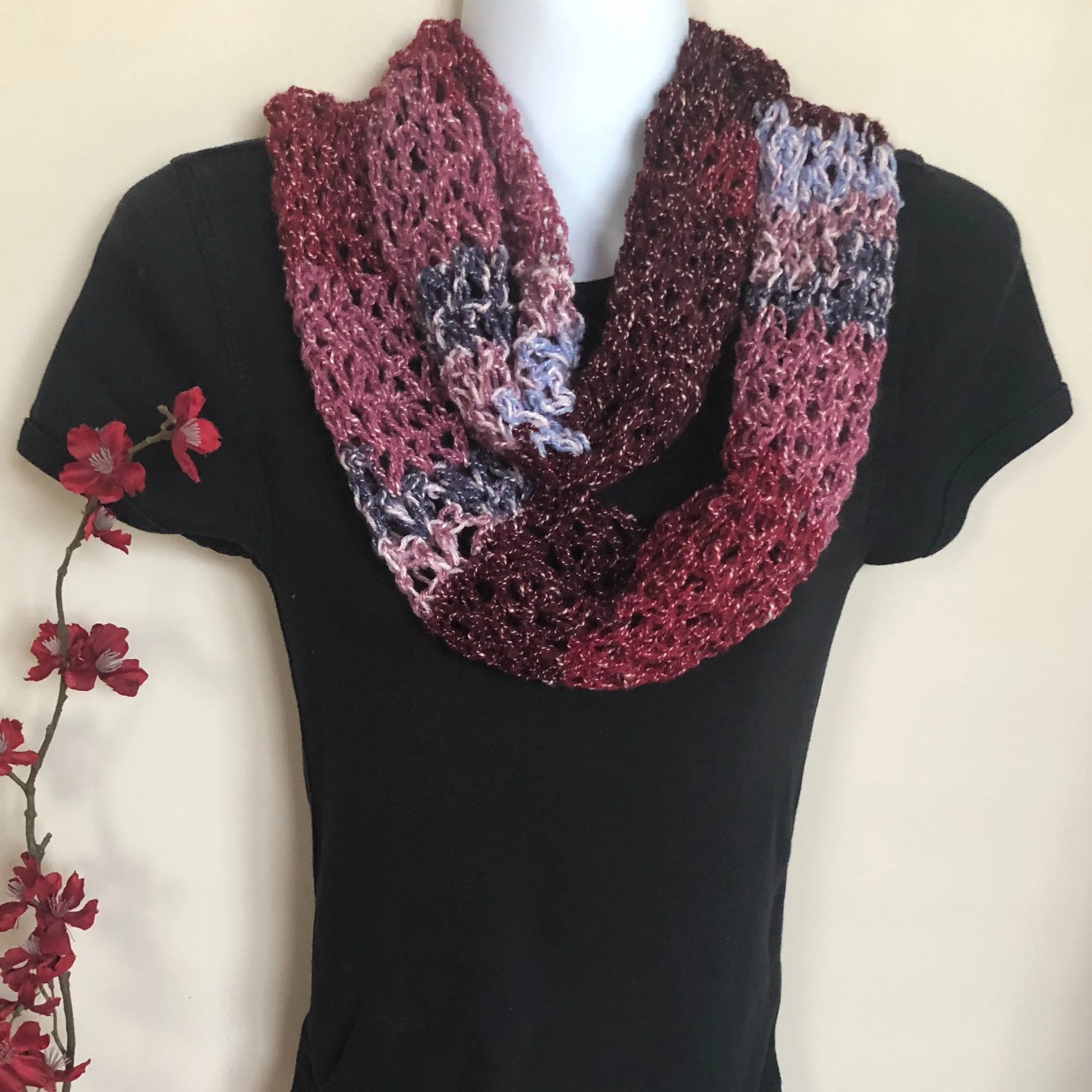 A mannequin wearing a multicolored crochet infinity scarf with shades of pink, purple, and dark red, paired with a black top with floral embroidery on the sleeve, next to a branch with pink flowers against a white background.