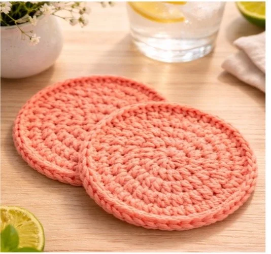 A pair of pink crocheted coasters on a wooden table, with a glass of water, a lime wedge, and white flowers in the background.
