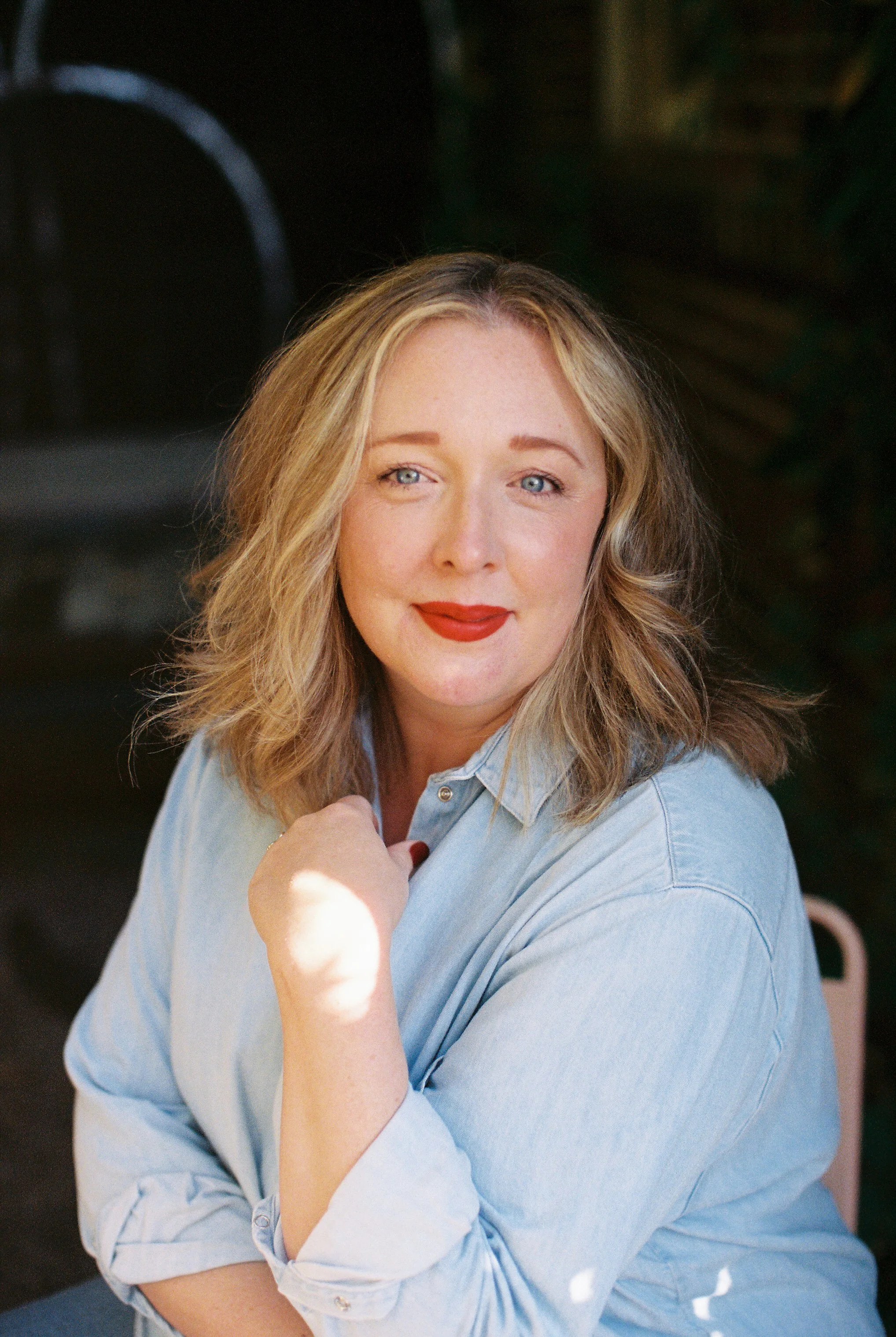 A photo fo Cate Croft. She has blue eyes and red lipstick, wearing a light blue button-up shirt, smiling softly at the camera with her chin resting on her hand.