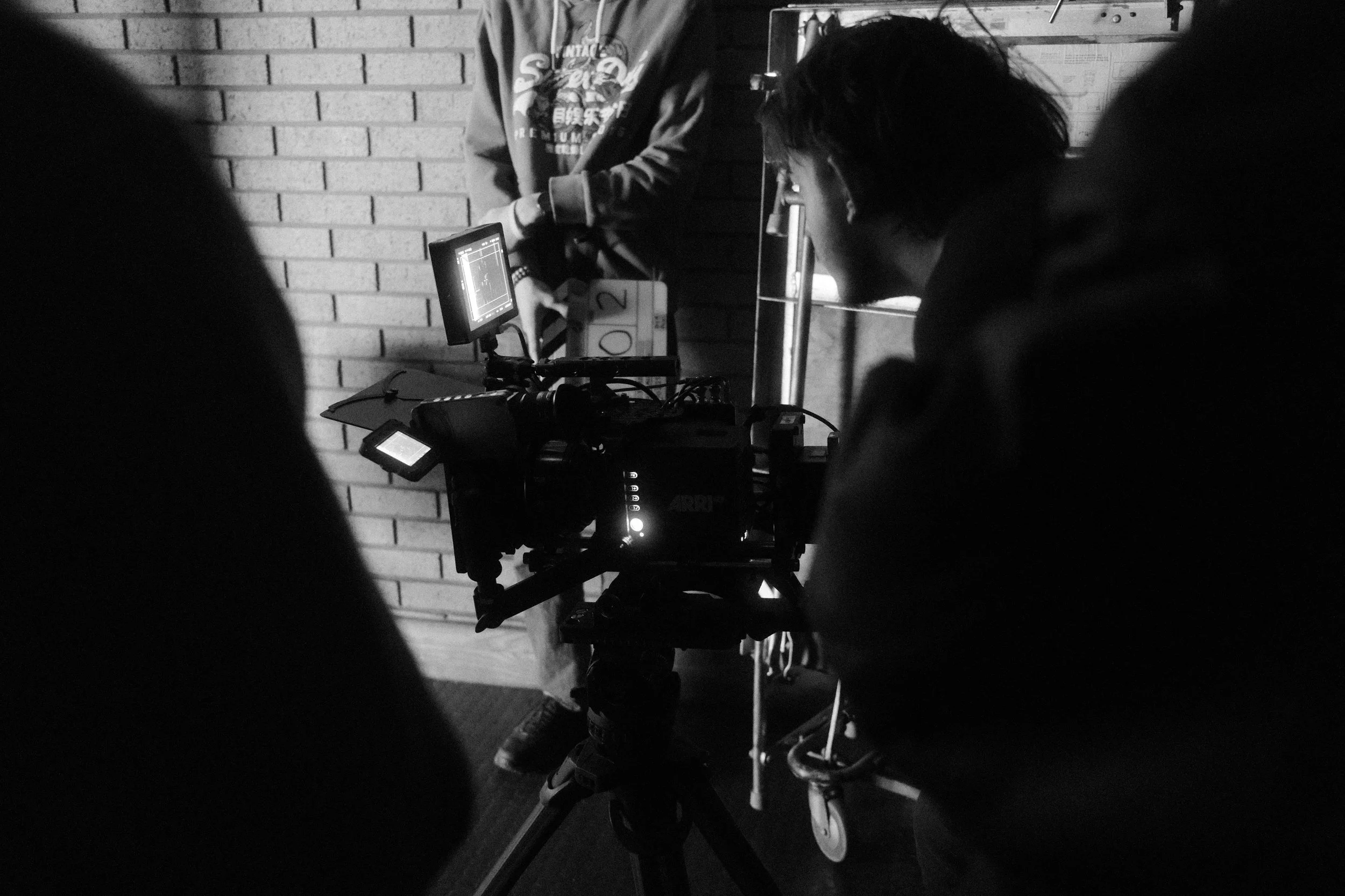 Black and white photo of people working with a professional film camera setup next to a brick wall.