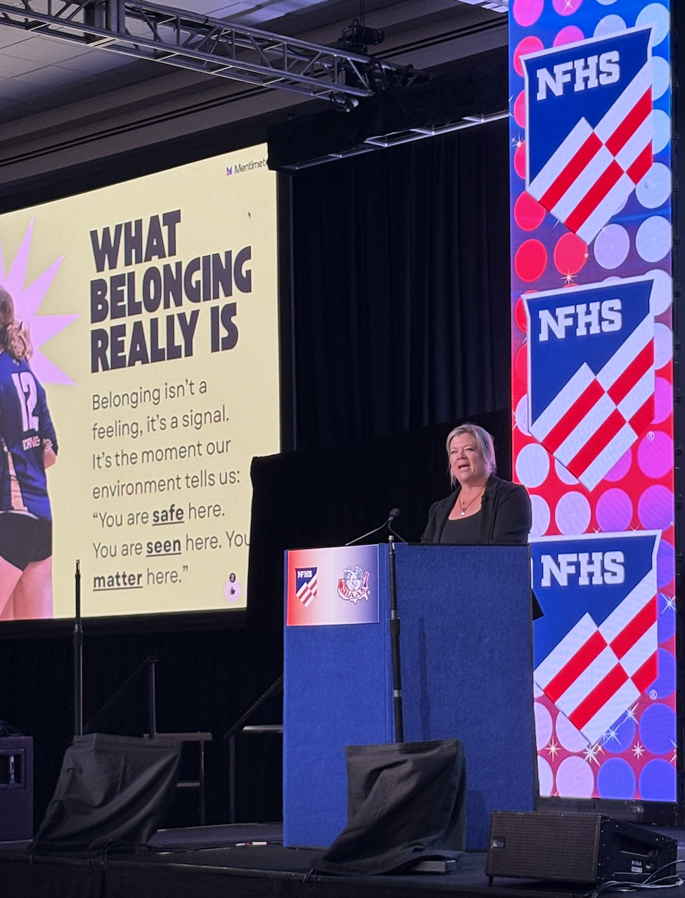 A woman speaking at a podium during a conference or event. Behind her is a large screen displaying a slide that reads "WHAT BELONGING REALLY IS" and explains the concept of belonging as a signal from the environment that communicates safety and being seen. The right side of the stage has vertical banners with the NFHS logo, which features a red and white striped shield with the letters NFHS.