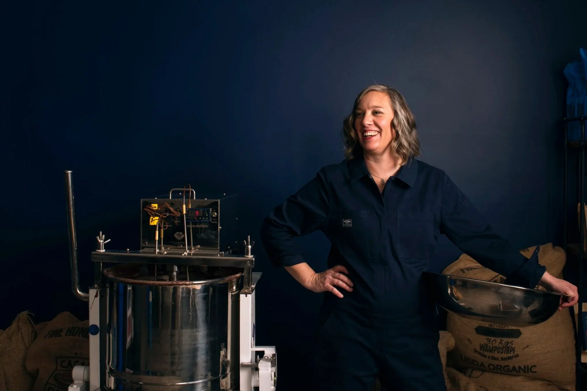 A woman with shoulder-length wavy hair wearing a navy blue jacket, smiling while standing next to a piece of scientific or technical equipment, with large sacks of organic material in the background, against a dark wall.