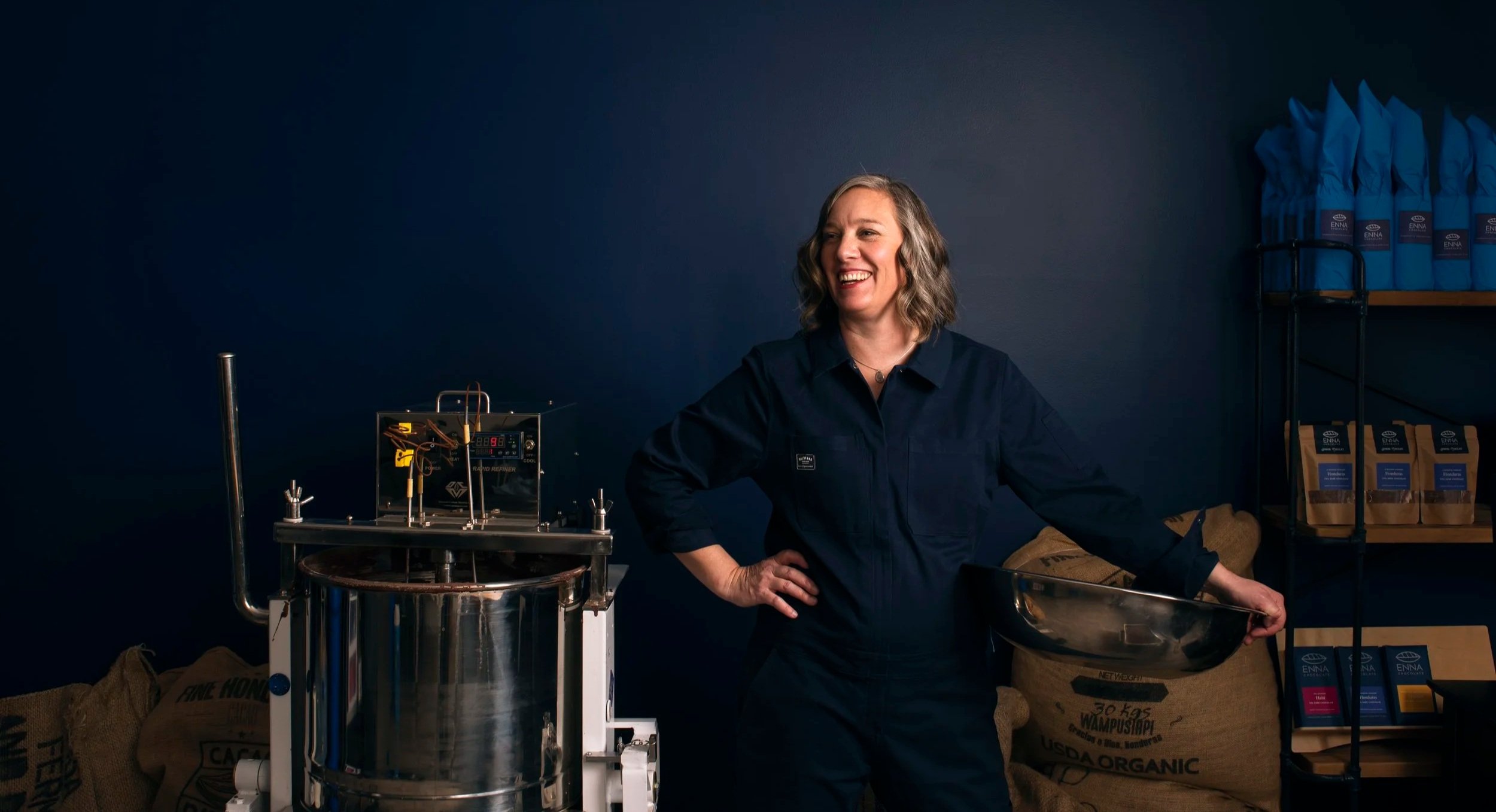 Woman with shoulder-length wavy hair smiling, wearing a dark blue shirt, standing in front of a blue wall, with scientific equipment and bags of organic material nearby.