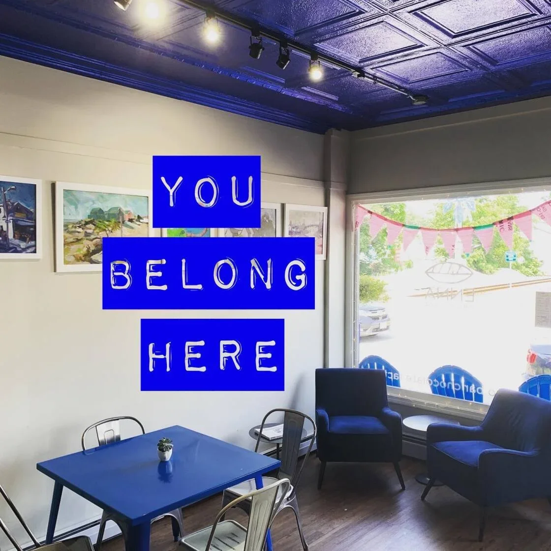 Inside Enna's cozy cafe with a blue ceiling, white walls with paintings, a window looking on the street outside, and blue furniture. Overlaid text reads 'YOU BELONG HERE' in blue letters.