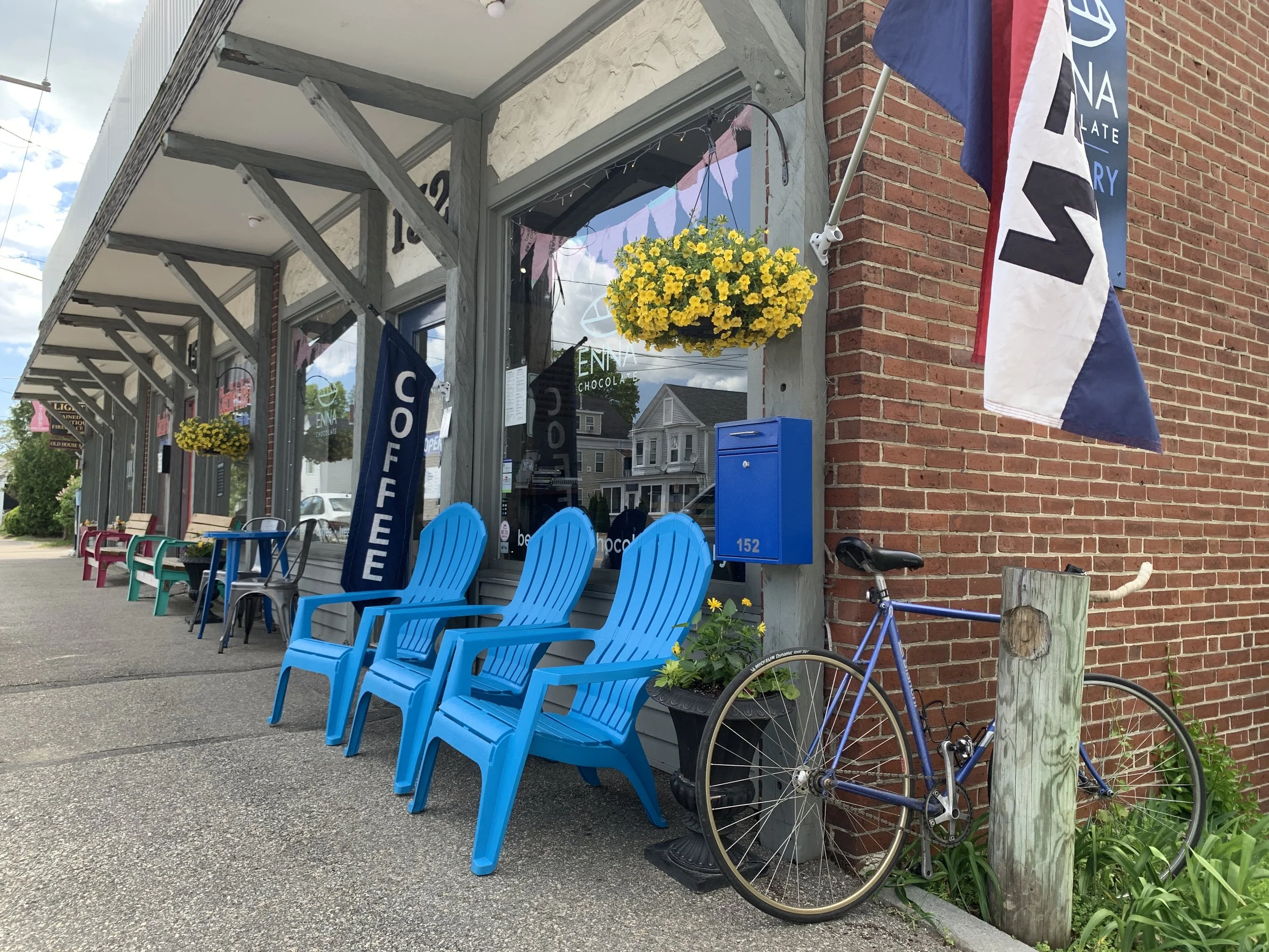 Exterior of a chocolate shop with blue Adirondack chairs outside, yellow flower hanging basket, flags, and a bicycle leaned against a wooden post.