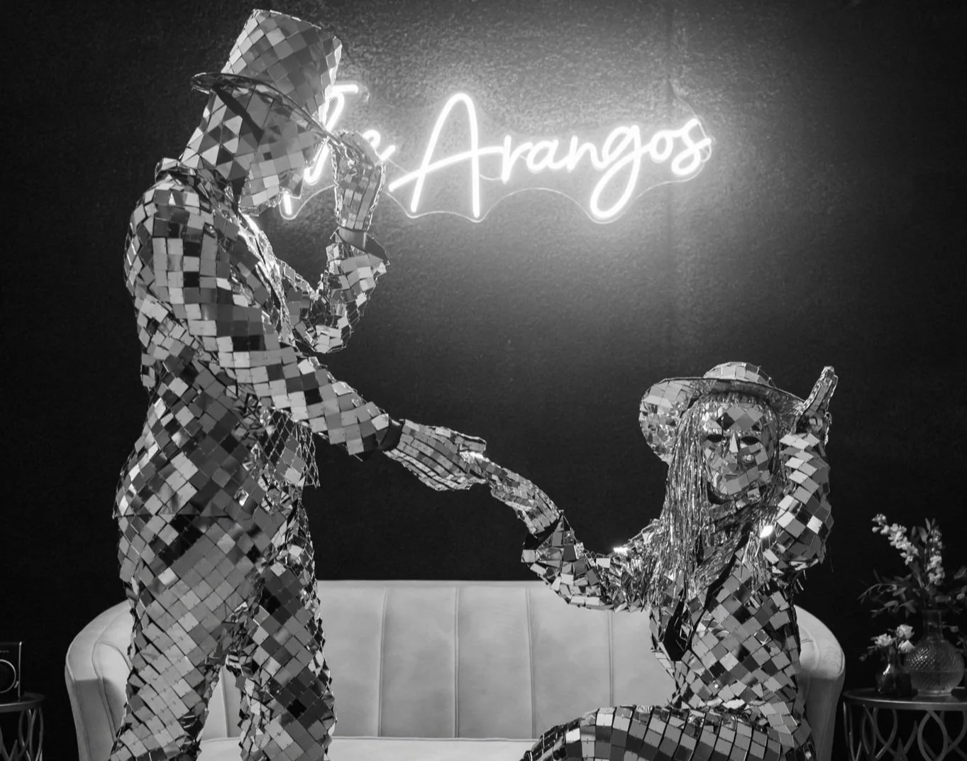 Two people dressed in mirrored disco outfits are in a room with a neon sign that reads 'The Arangos.' One person is standing, holding the other person's hand and has a geometric disco hat. The other person is kneeling with long hair and a headband, holding a phone. There are decorative plants and flowers nearby.