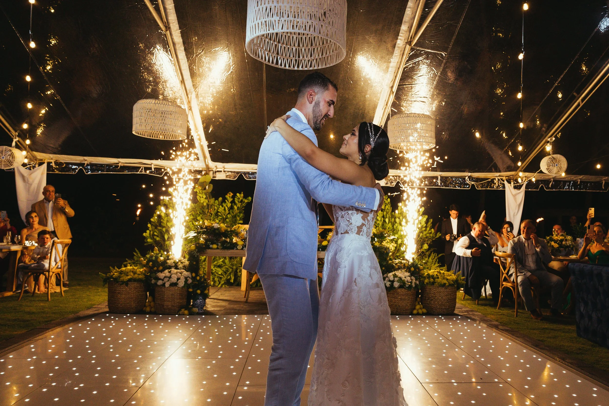 A bride and groom dancing at their wedding reception under a decorated tent with string lights and fireworks in the background, with guests watching and celebrating.