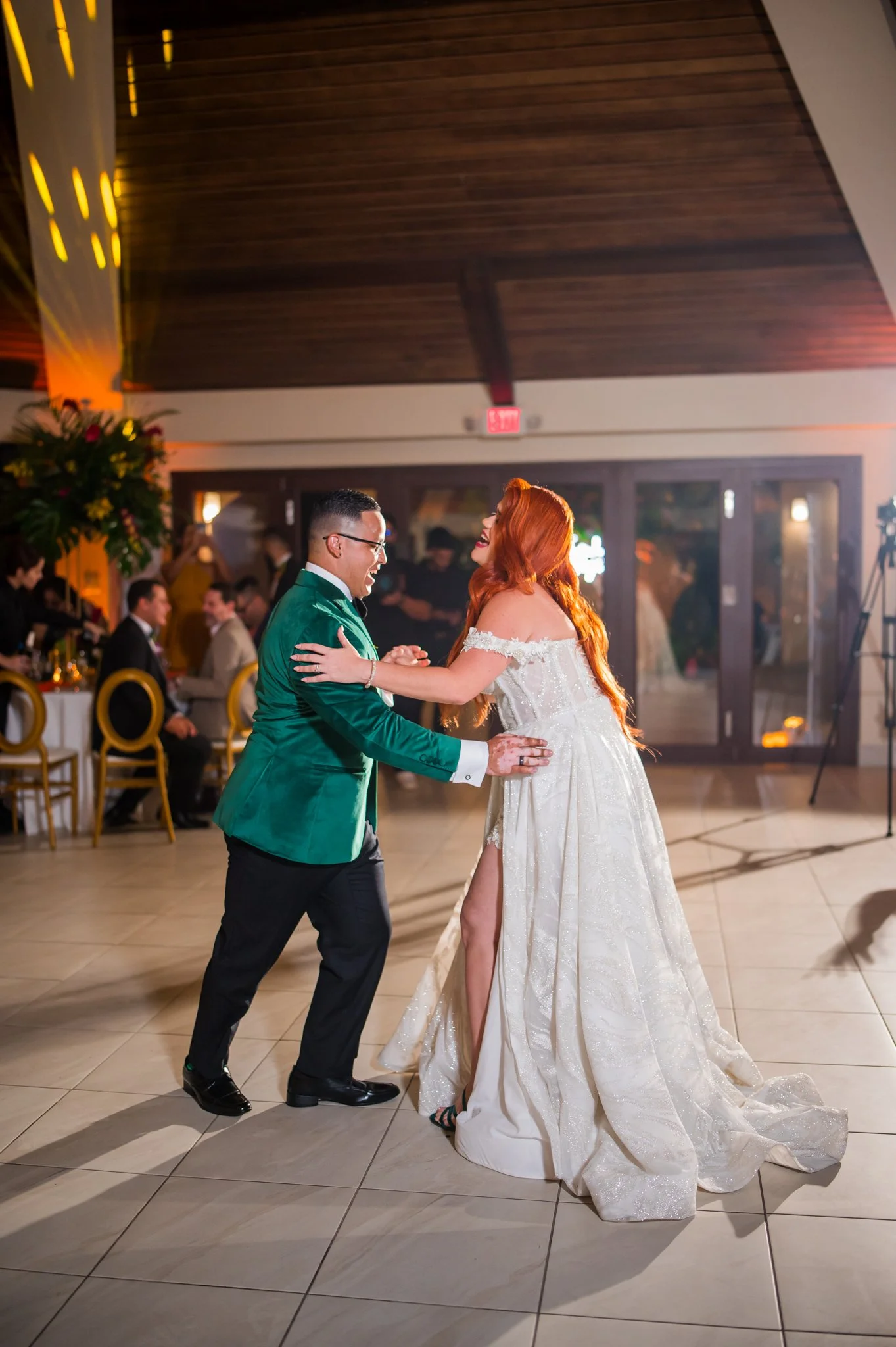 A bride and groom dancing at their wedding reception, with guests seated in the background and warm lighting in the venue.