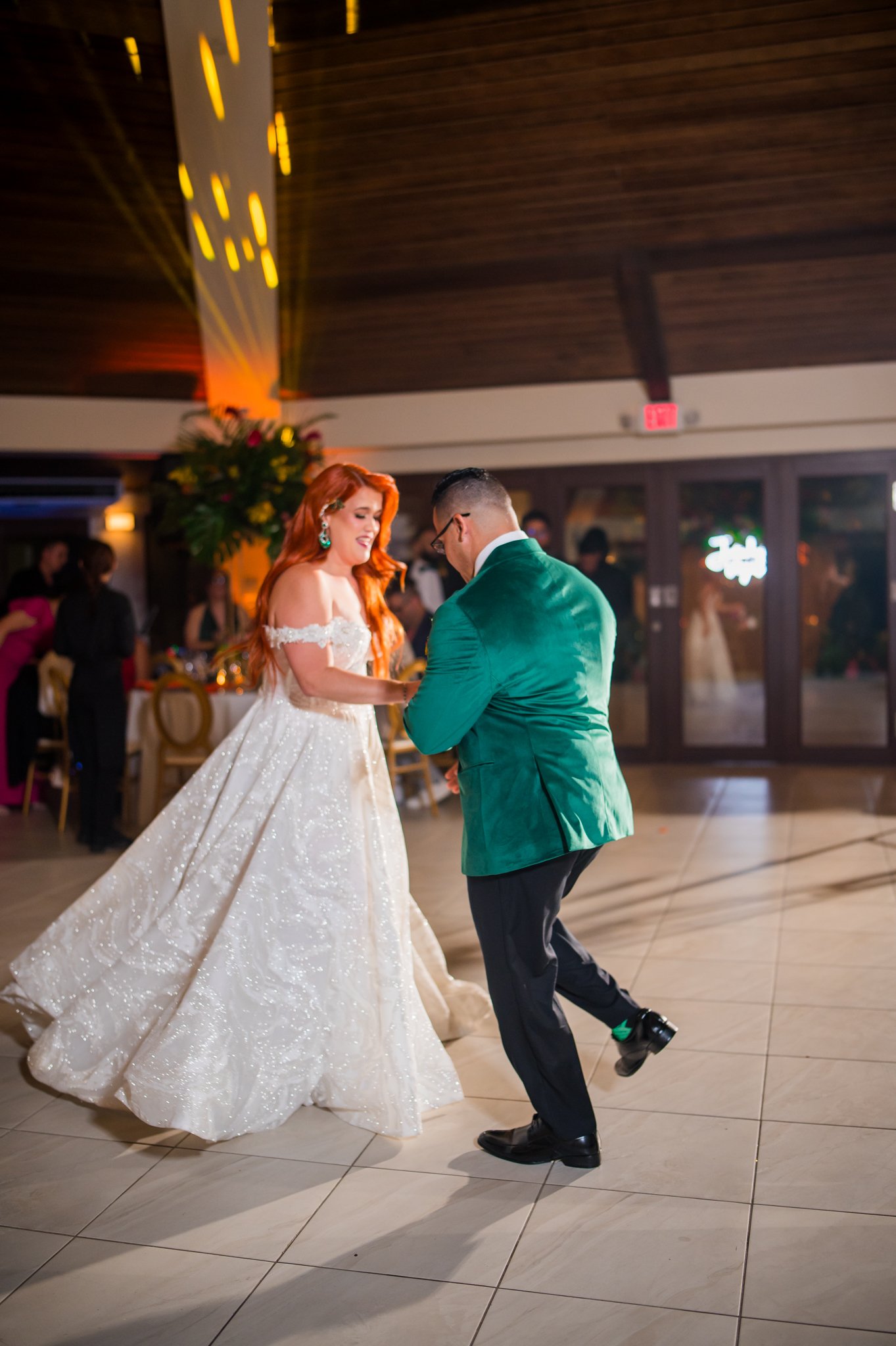 A bride with red hair in a white dress dancing with a groom in a green jacket at a wedding reception.