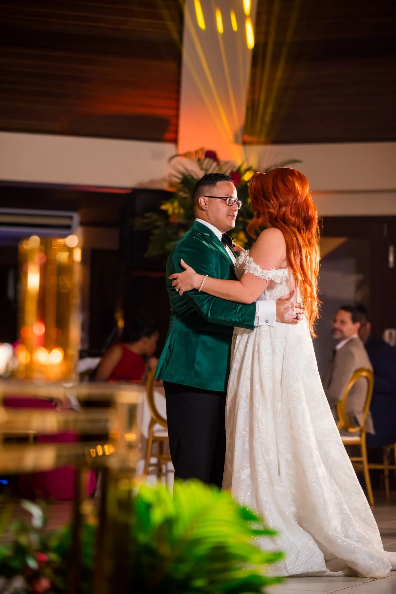 A couple dancing at a wedding reception, the man in a green velvet tuxedo and the woman in a white wedding gown, with guests sitting in the background.