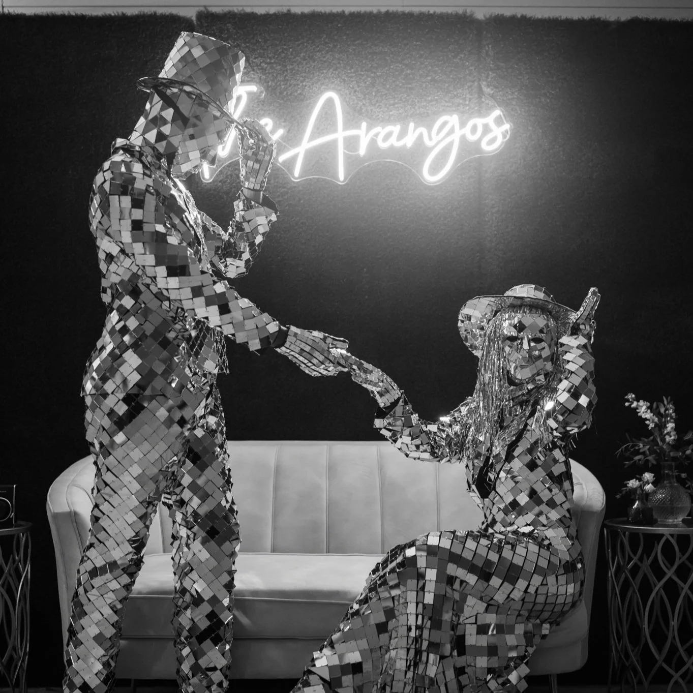 Two people wearing full-body mirror mosaic suits, one kneeling and the other standing, in front of a neon sign that says 'The Arangos', with a sofa and floral arrangement nearby.