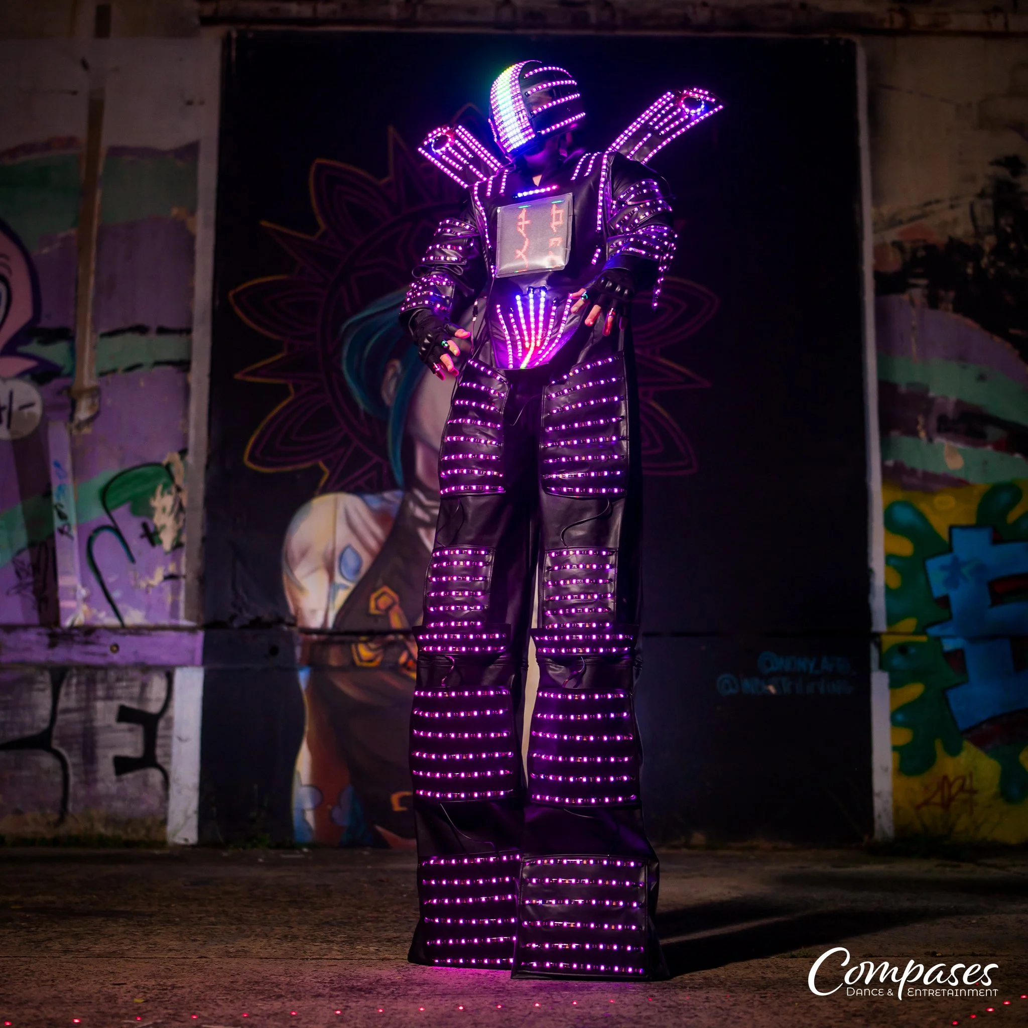 A person in a futuristic outfit covered in purple LED lights standing on a dark street in front of graffiti art, with the name 'Compases Dance & Entertainment' in the bottom right corner.