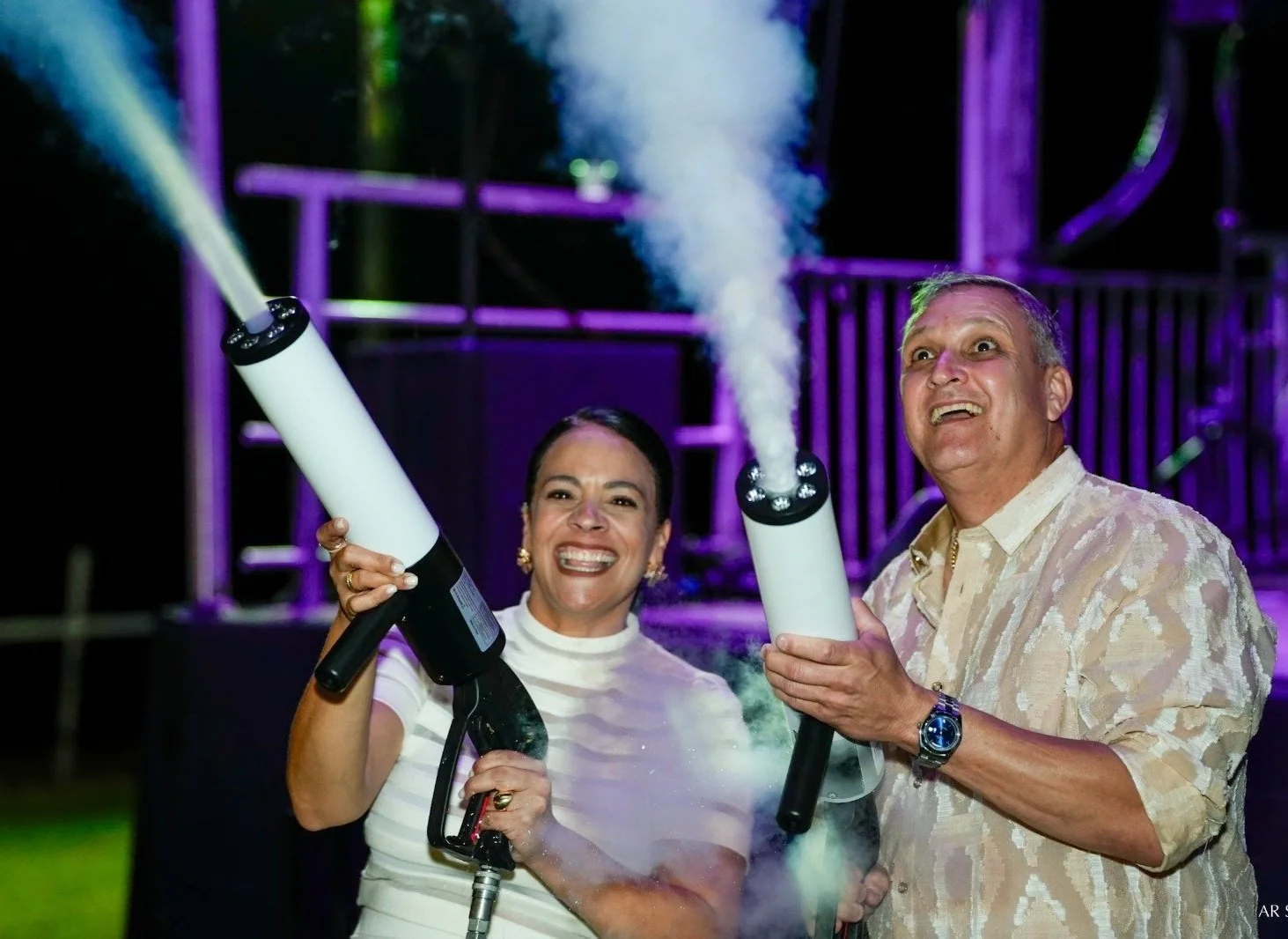 Two adults, a woman and a man, using smoke or fog machines at night, with colorful lighting in the background.