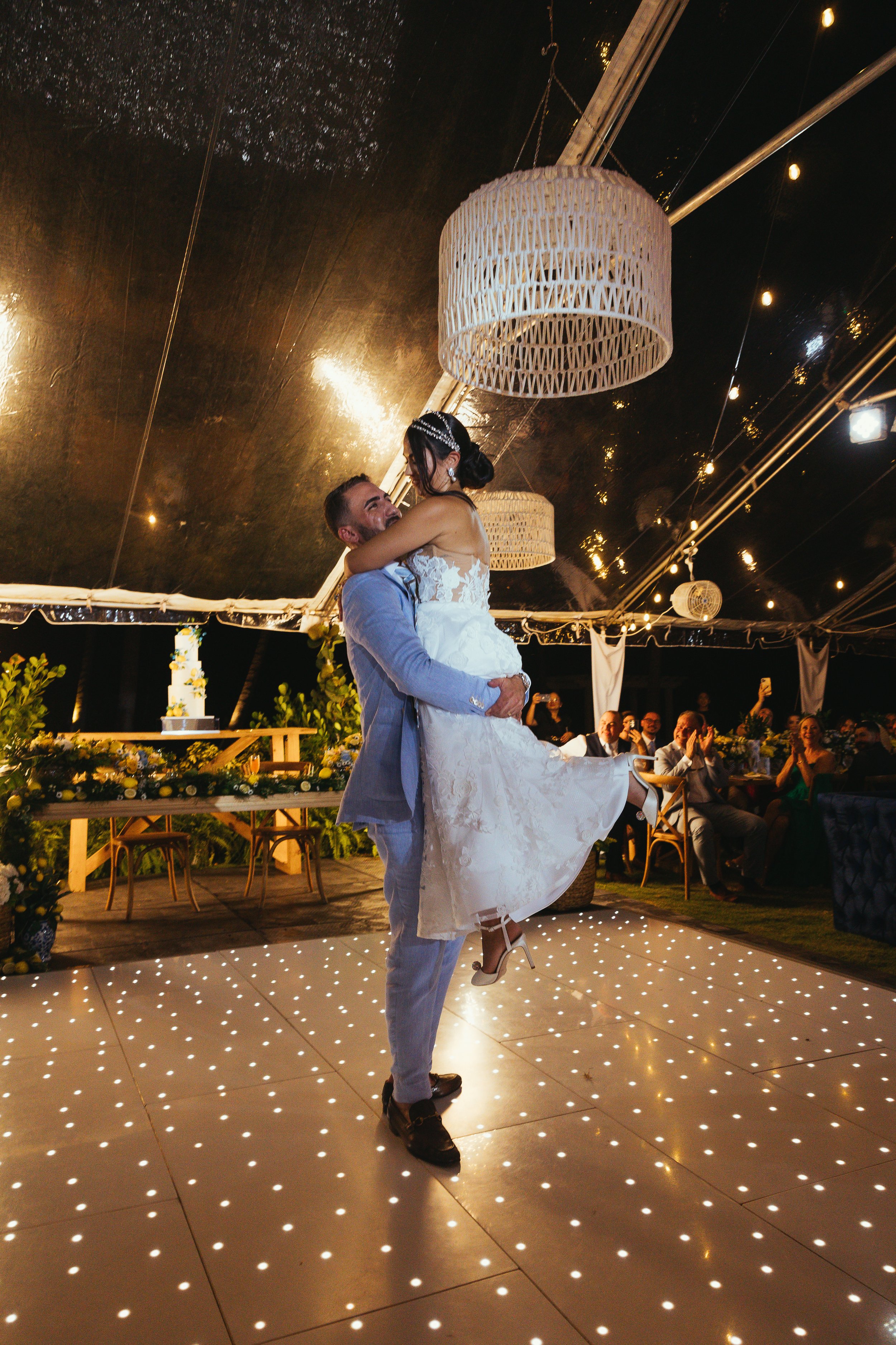 A couple dancing at their wedding reception, with guests clapping and smiling in the background.