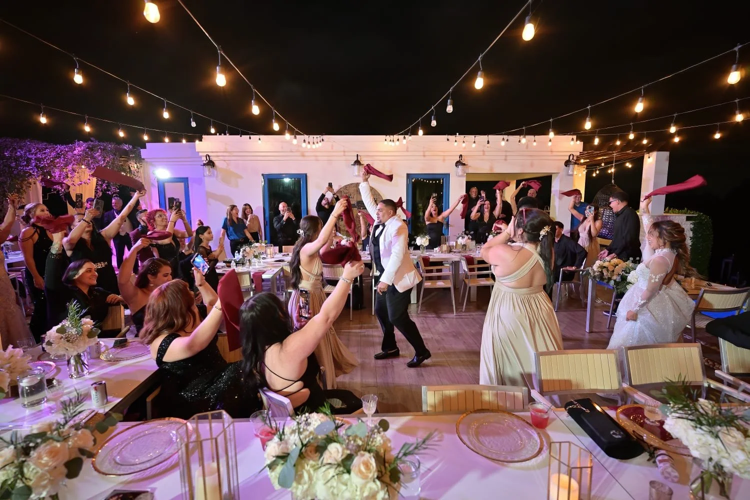 A wedding reception with guests dancing and taking photos outdoors at night, with string lights overhead and decorated tables.