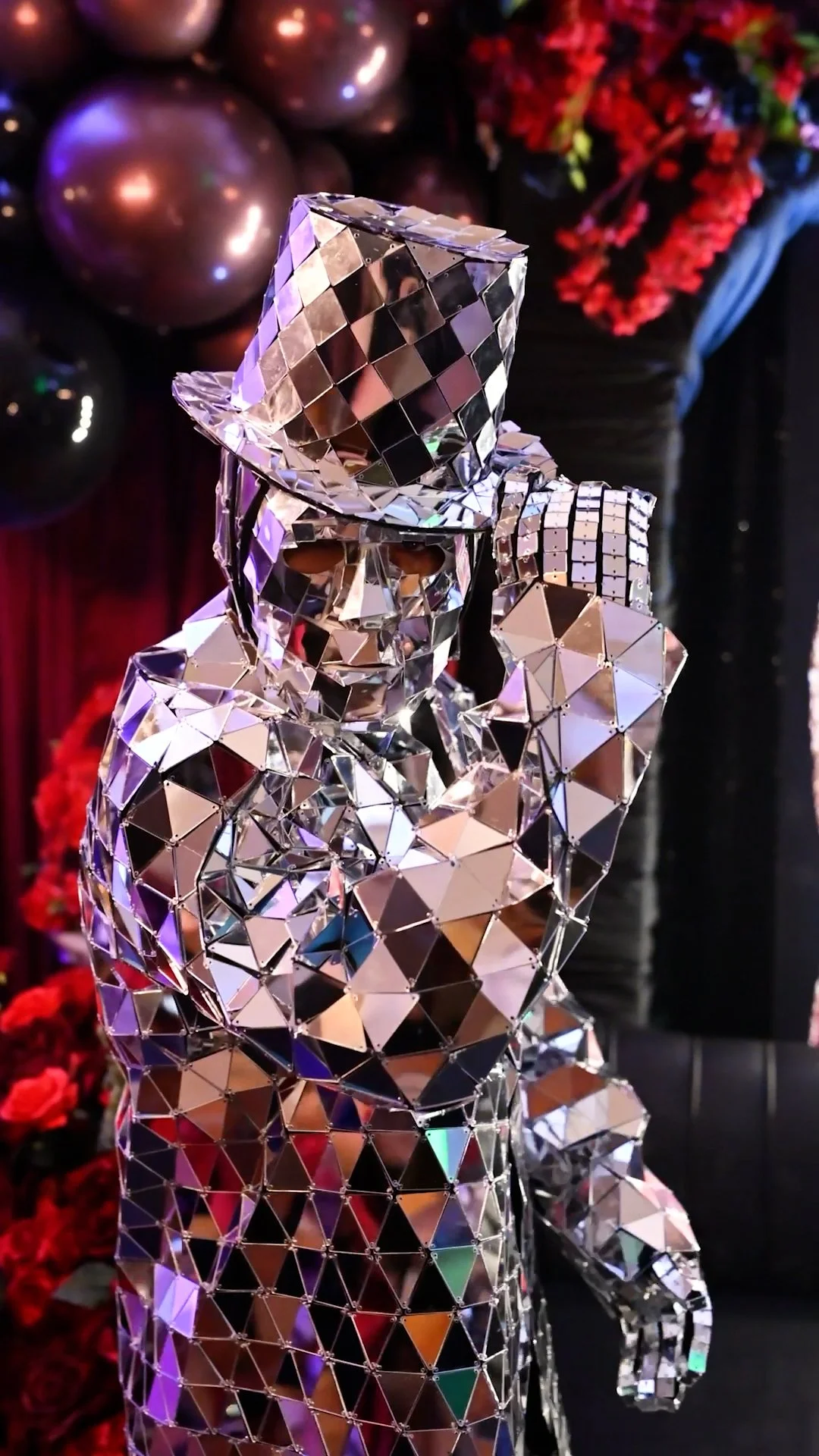 A mirror mosaic sculpture of a person wearing a hat and glasses, with a hand raised to their face, reflected in the shiny surfaces.