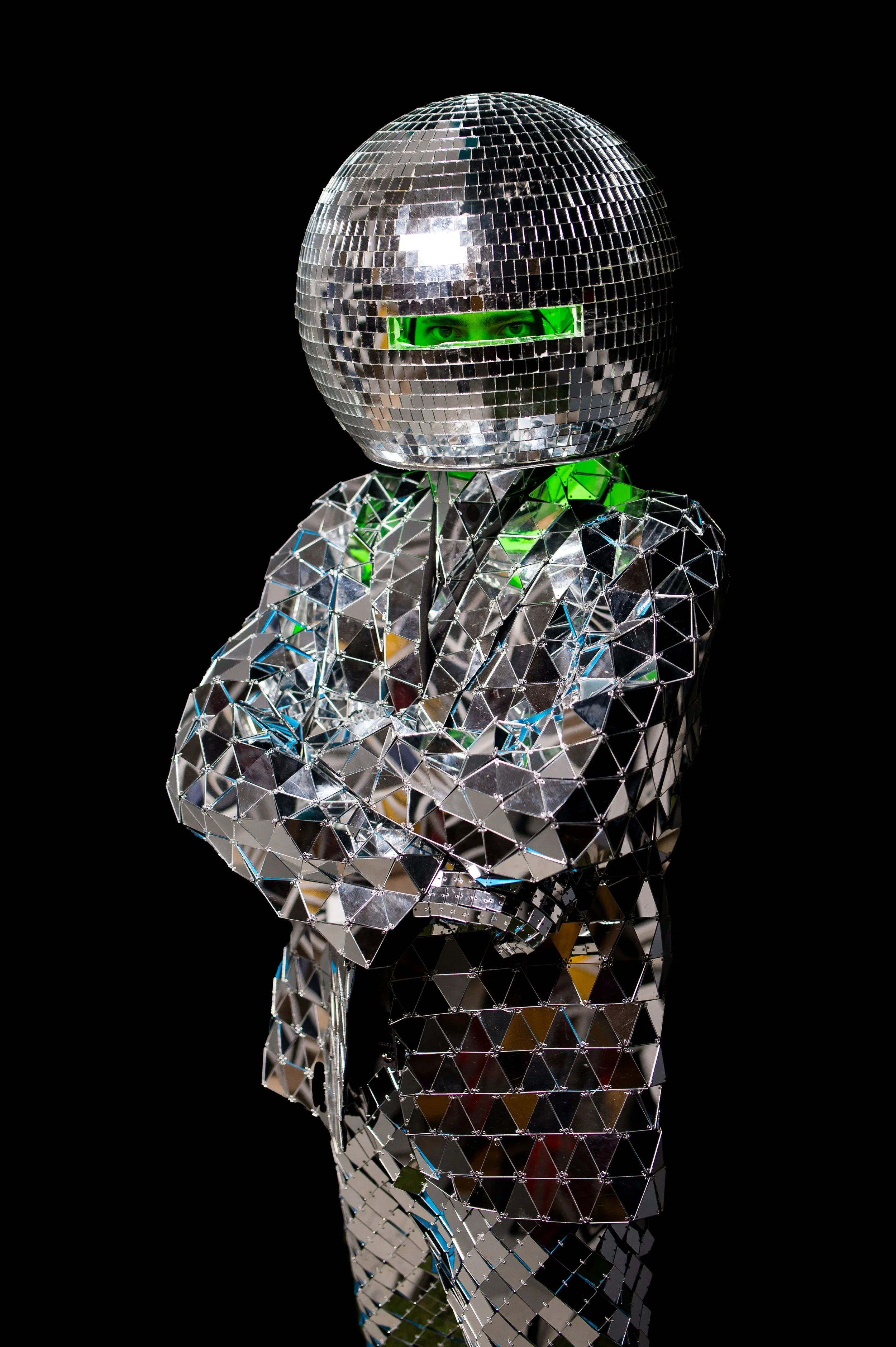 Person in a silver mirrored, geometrically patterned suit and helmet resembling a disco ball, with reflective surfaces and a green visor, standing with arms crossed against a black background.