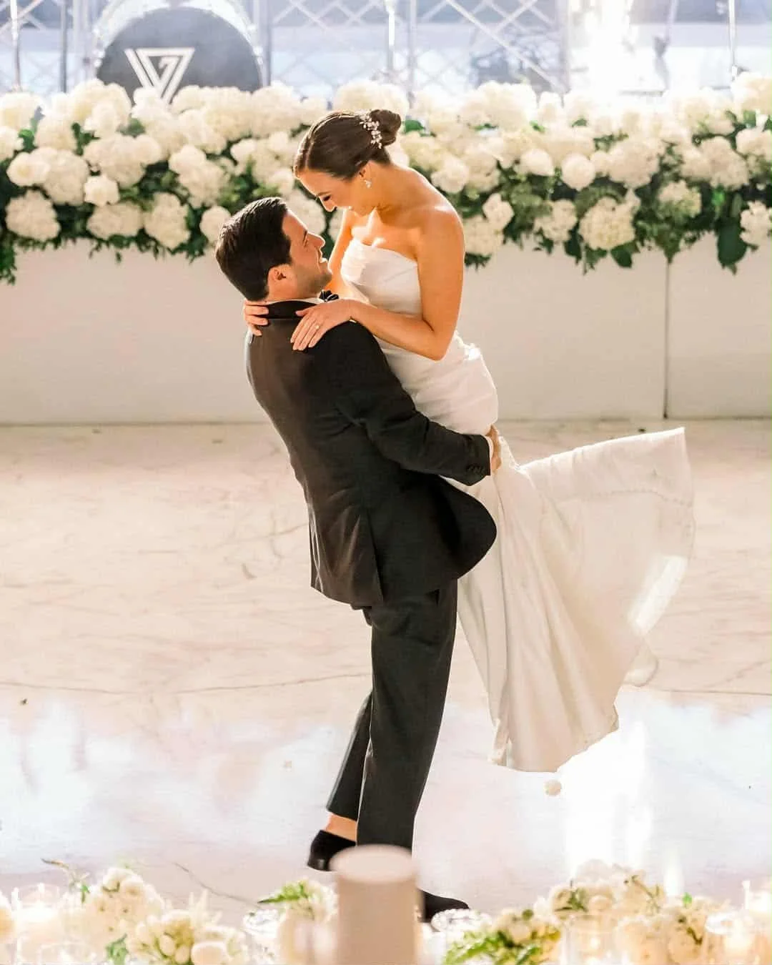 A bride and groom dancing at their wedding reception, with the groom lifting the bride in his arms and both smiling joyfully, surrounded by floral decorations.