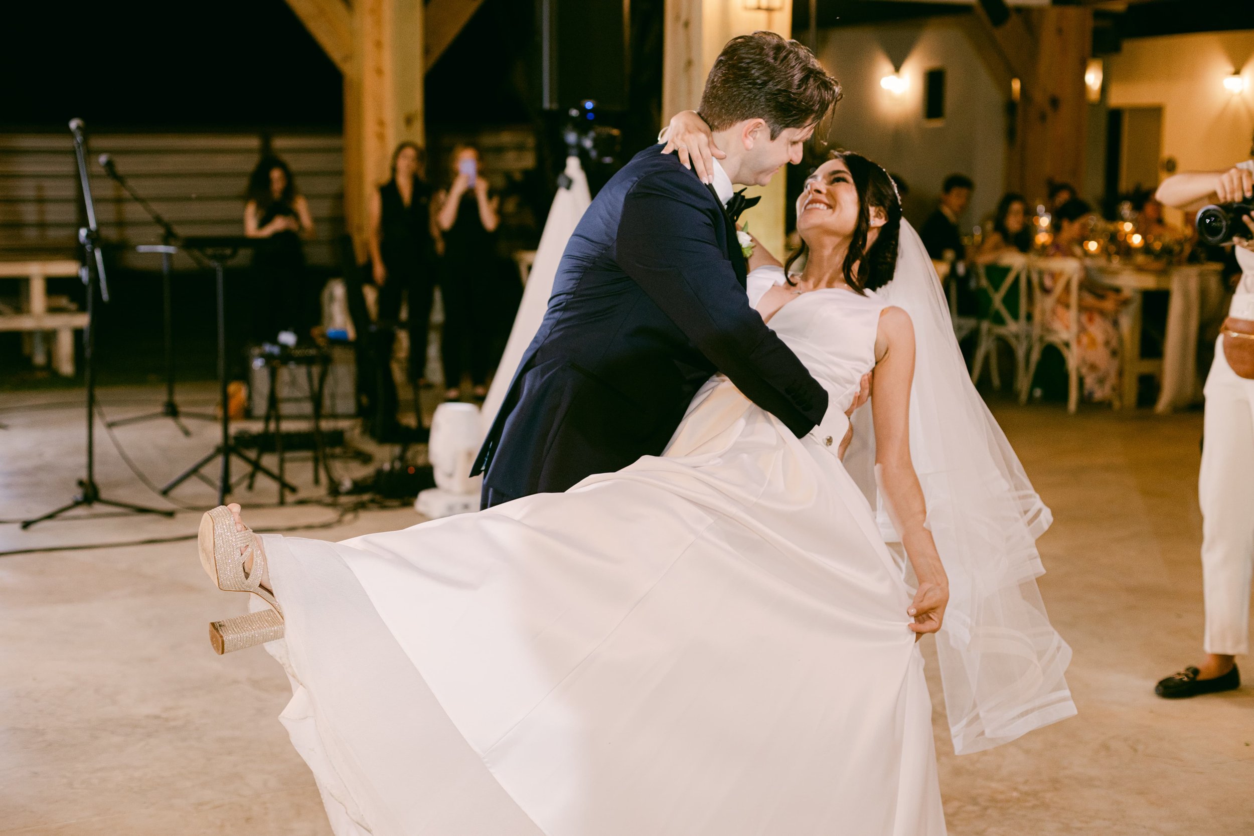 A groom in a tuxedo dips and twirls a bride in a white wedding dress during a dance at a wedding reception, with guests and a photographer in the background.