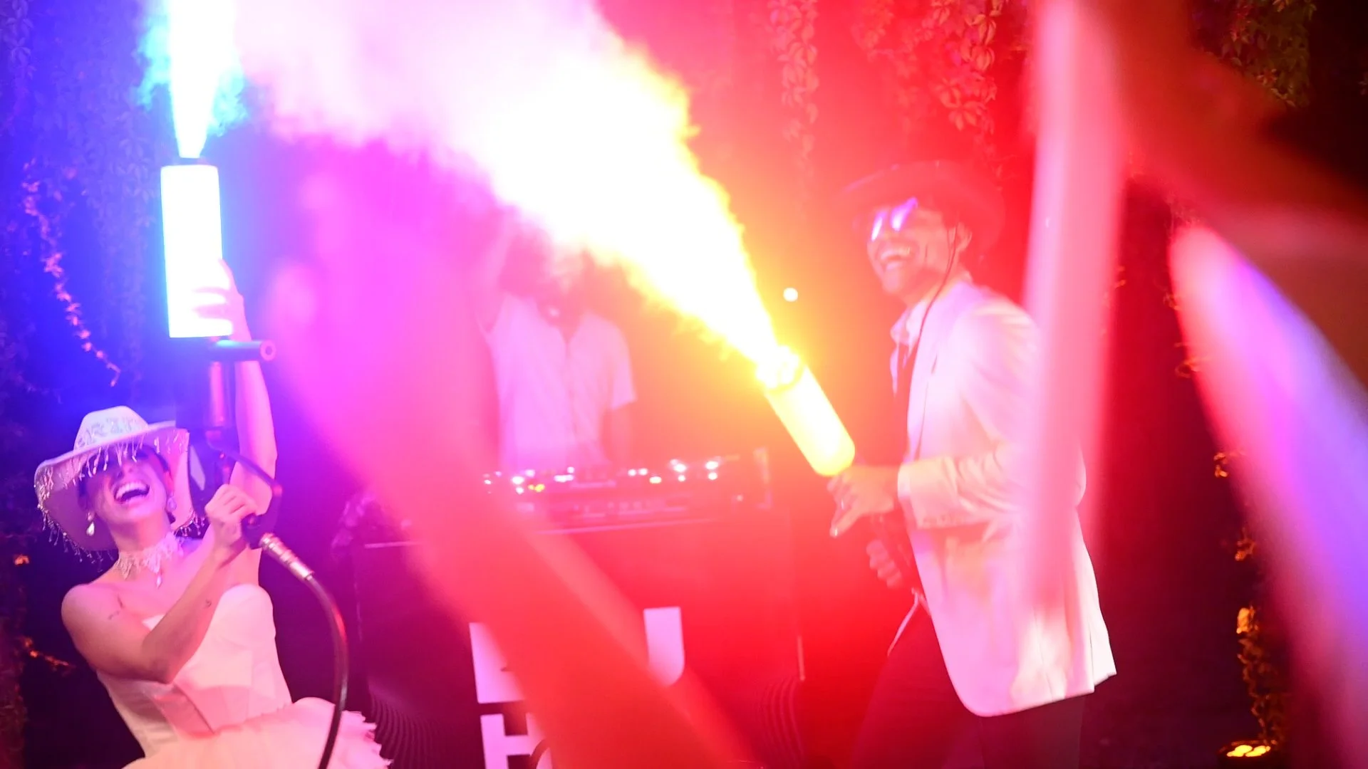 Two people smiling and playing with fog machines at night, with colorful stage lights in the background.