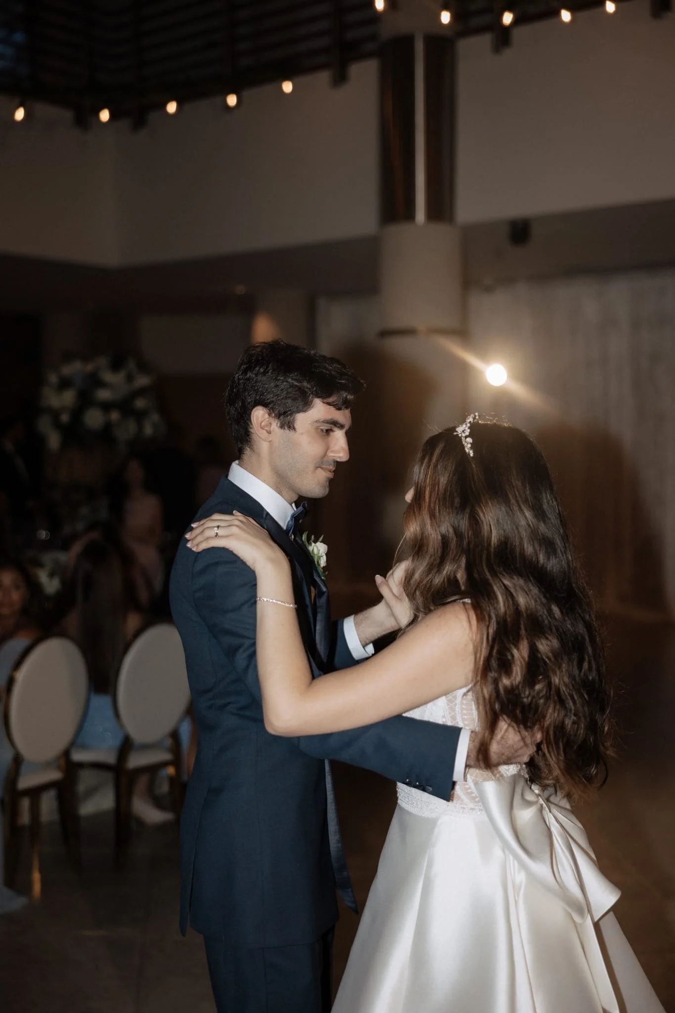A couple dancing at a wedding reception, with the groom in a navy suit and the bride in a white dress, dancing closely together in a decorated event hall.