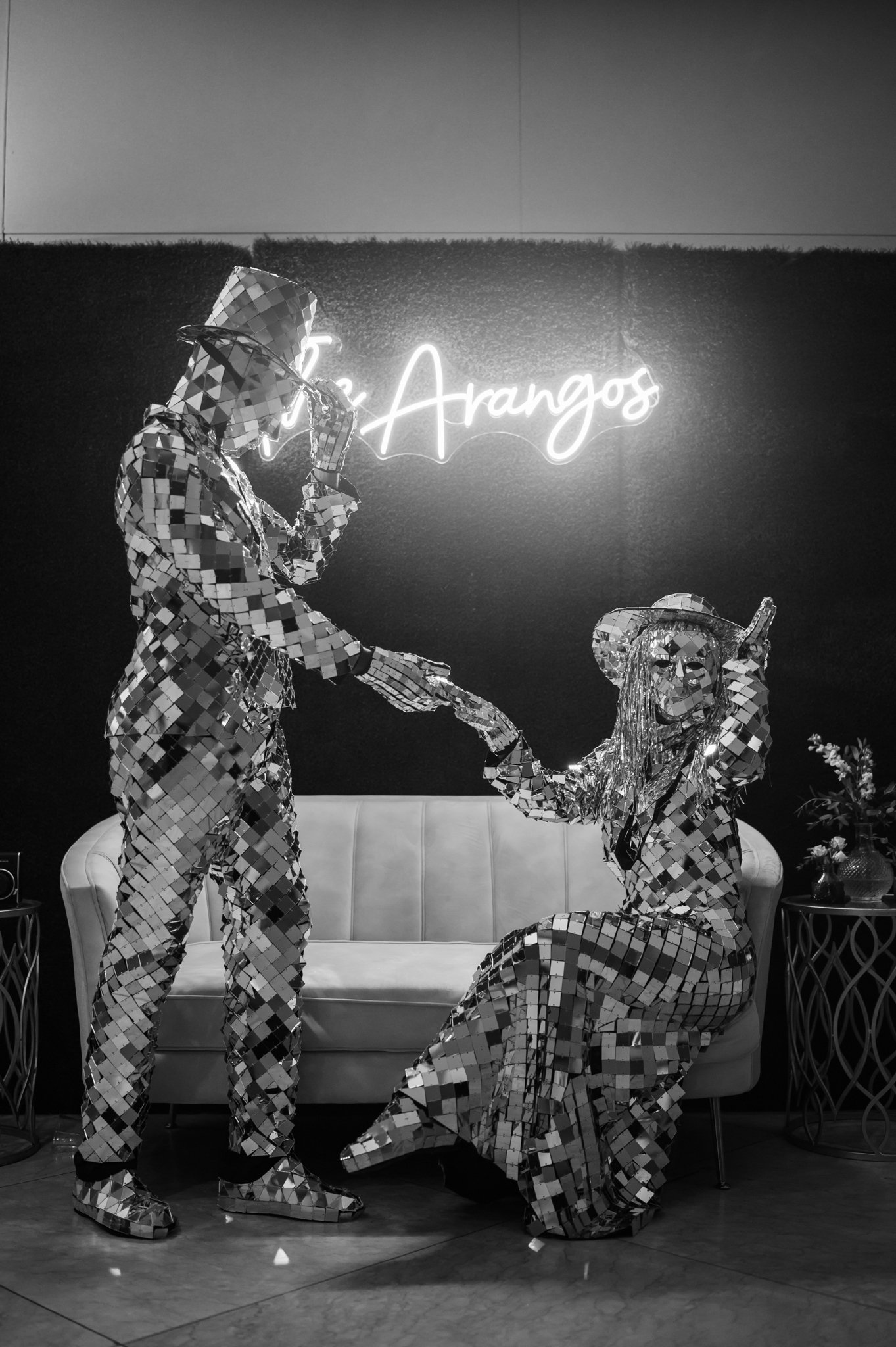 Two individuals dressed in reflective, mirror-like suits and accessories, sitting and standing on a vintage-style sofa in a modern room with a neon sign that reads 'Los Arangos' behind them.