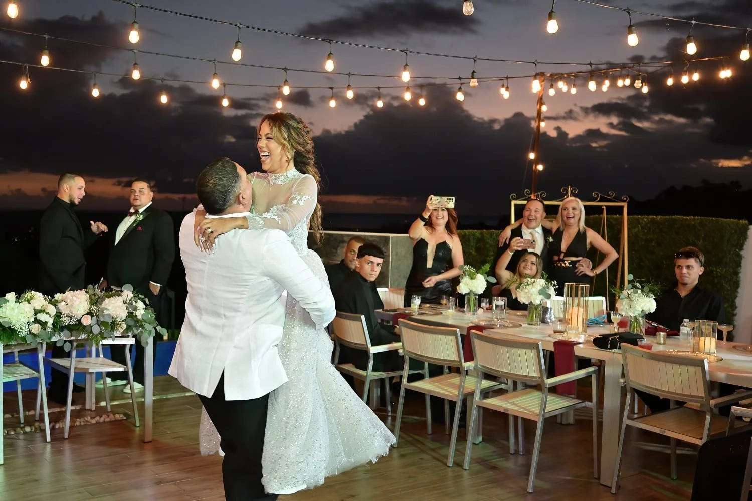 People at a wedding reception celebrating, with a woman in a wedding dress being lifted by a man, while others watch and take photos, under string lights at sunset.
